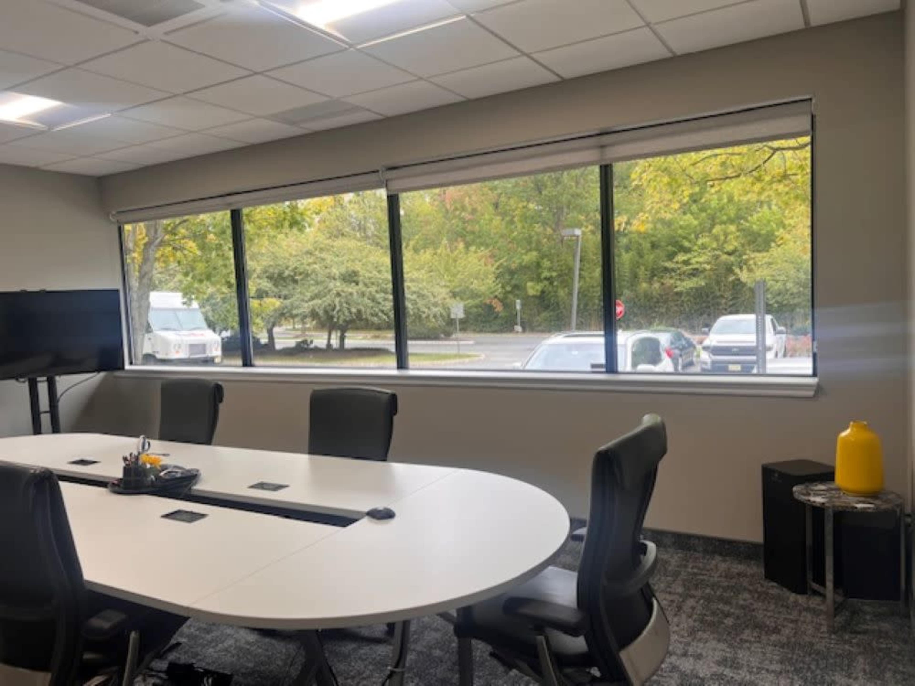 A conference room features a large round table with chairs, facing a window that overlooks a tree-lined parking lot.