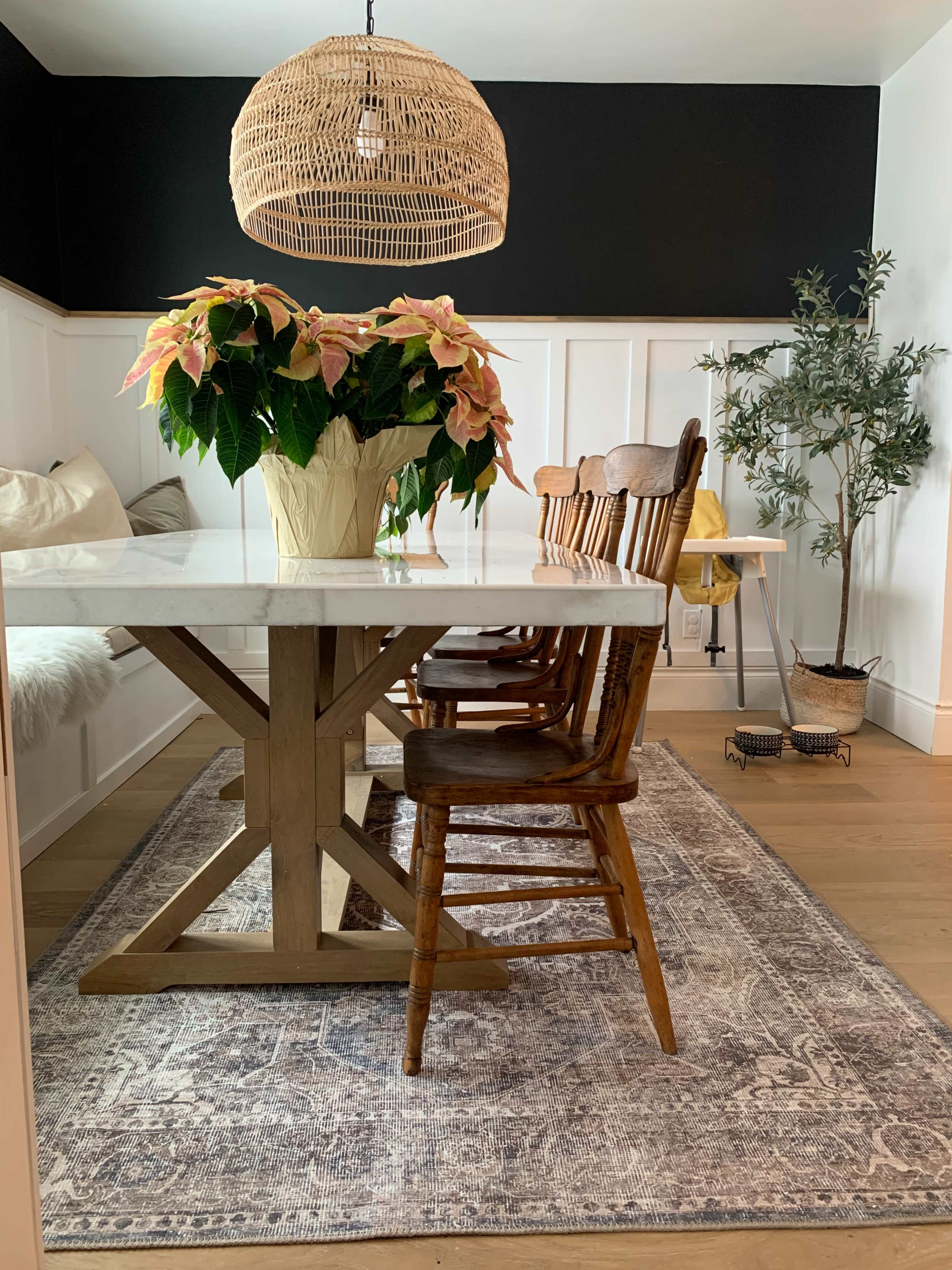 A modern dining room features a large wooden table surrounded by several chairs, with a potted plant in the center and a rattan pendant light overhead.