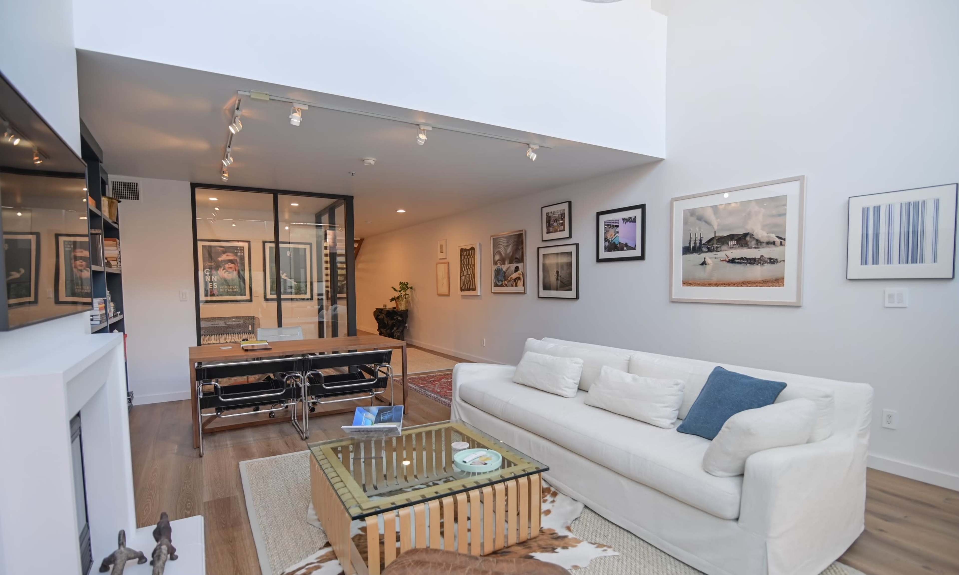 The image shows a bright, modern living space featuring a white sofa, a wooden table, and various framed artworks on the walls.