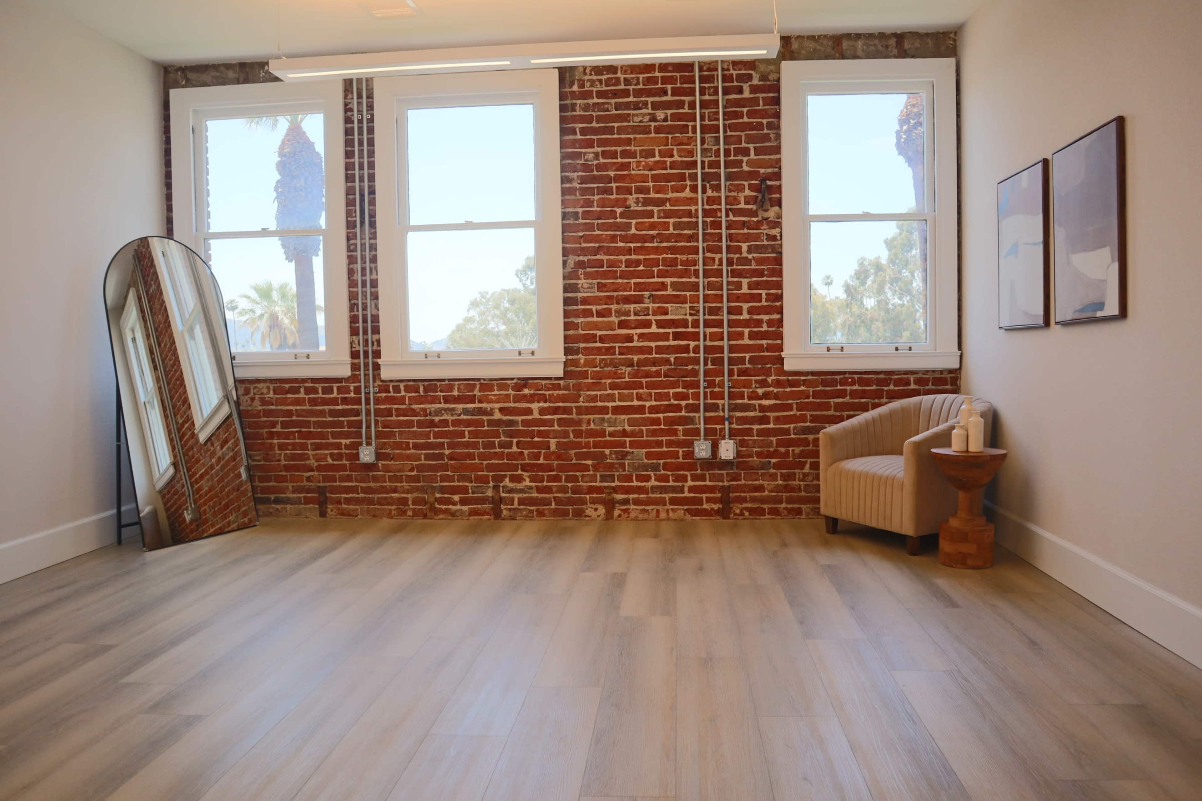 A minimalist interior with a curved mirror, a beige chair, and exposed brick walls near large windows.