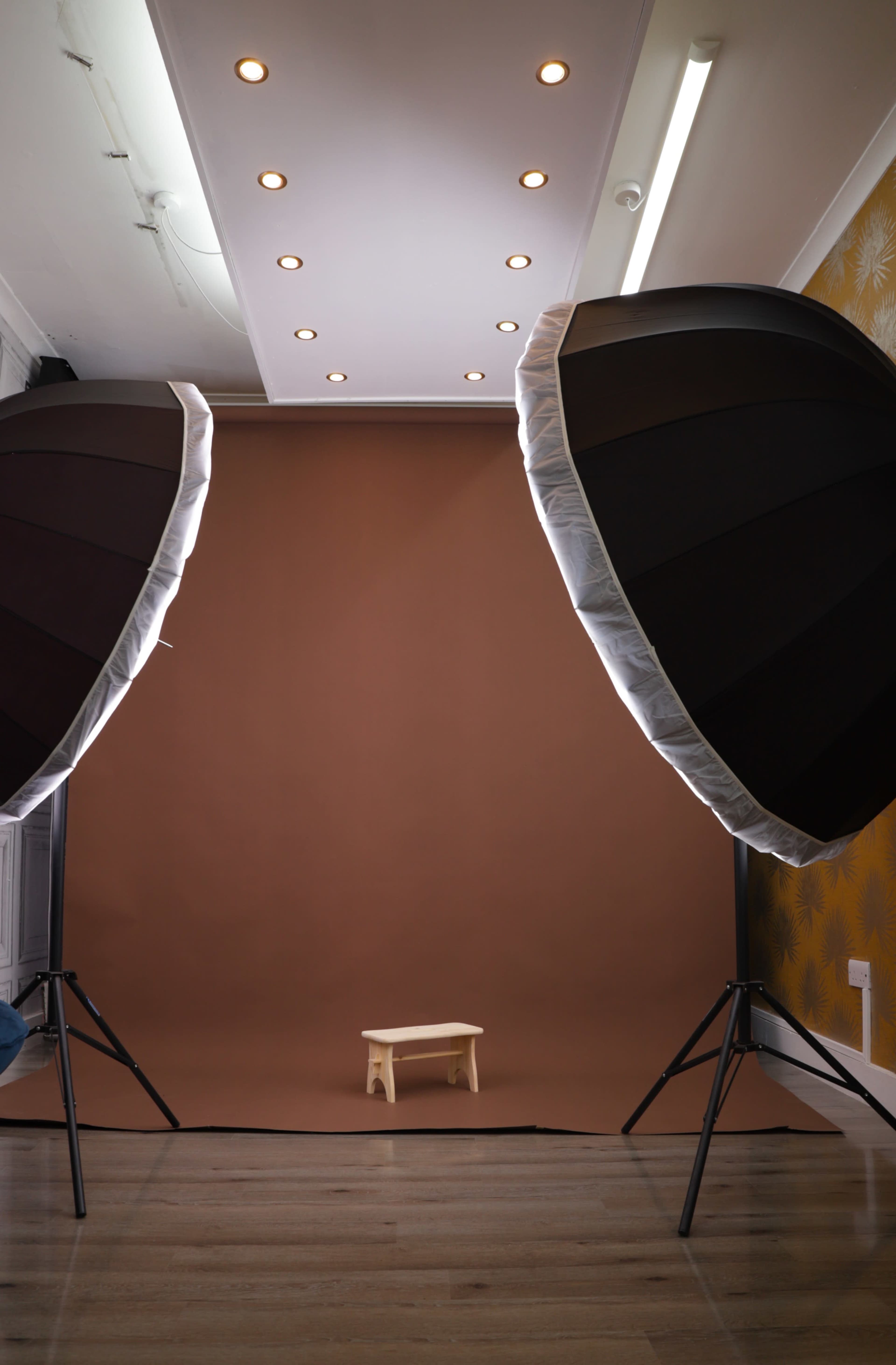 Two large softbox lights flank a small wooden stool in front of a brown backdrop inside a photography studio.
