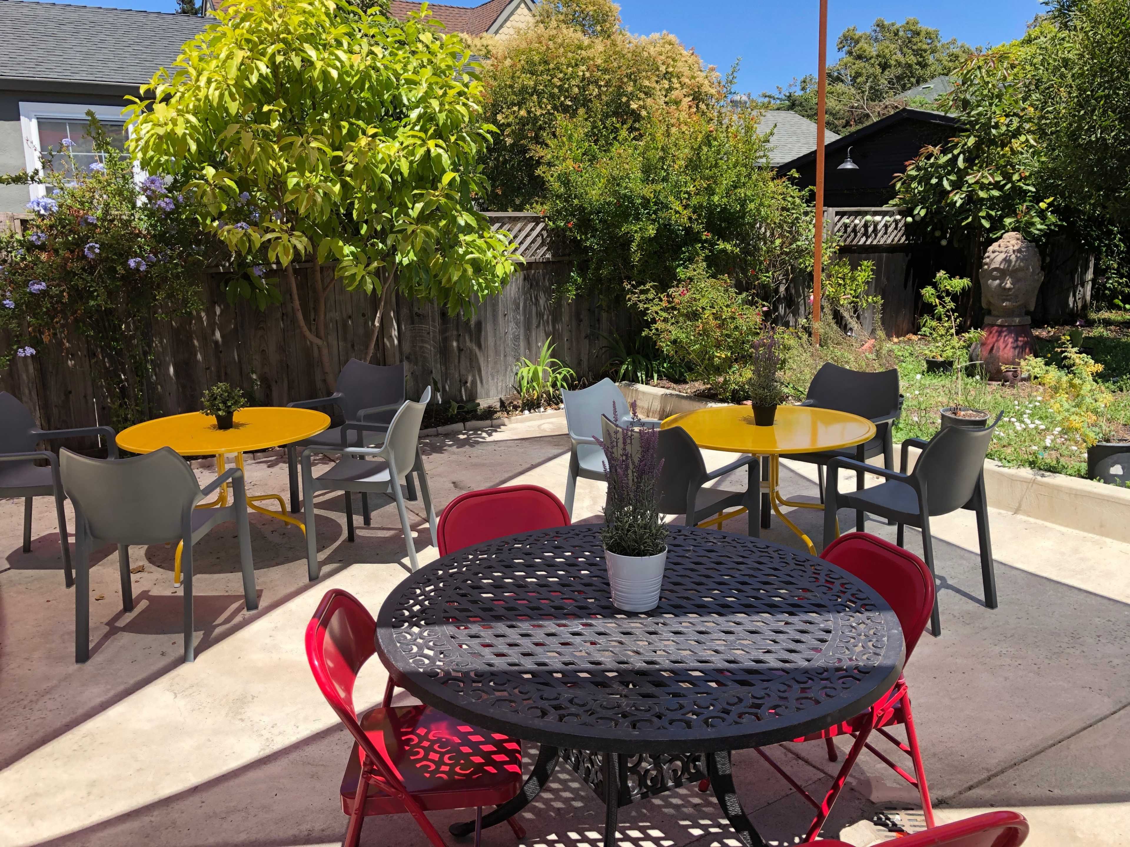 A outdoor seating area features several tables in yellow and black, surrounded by greenery and colorful chairs.