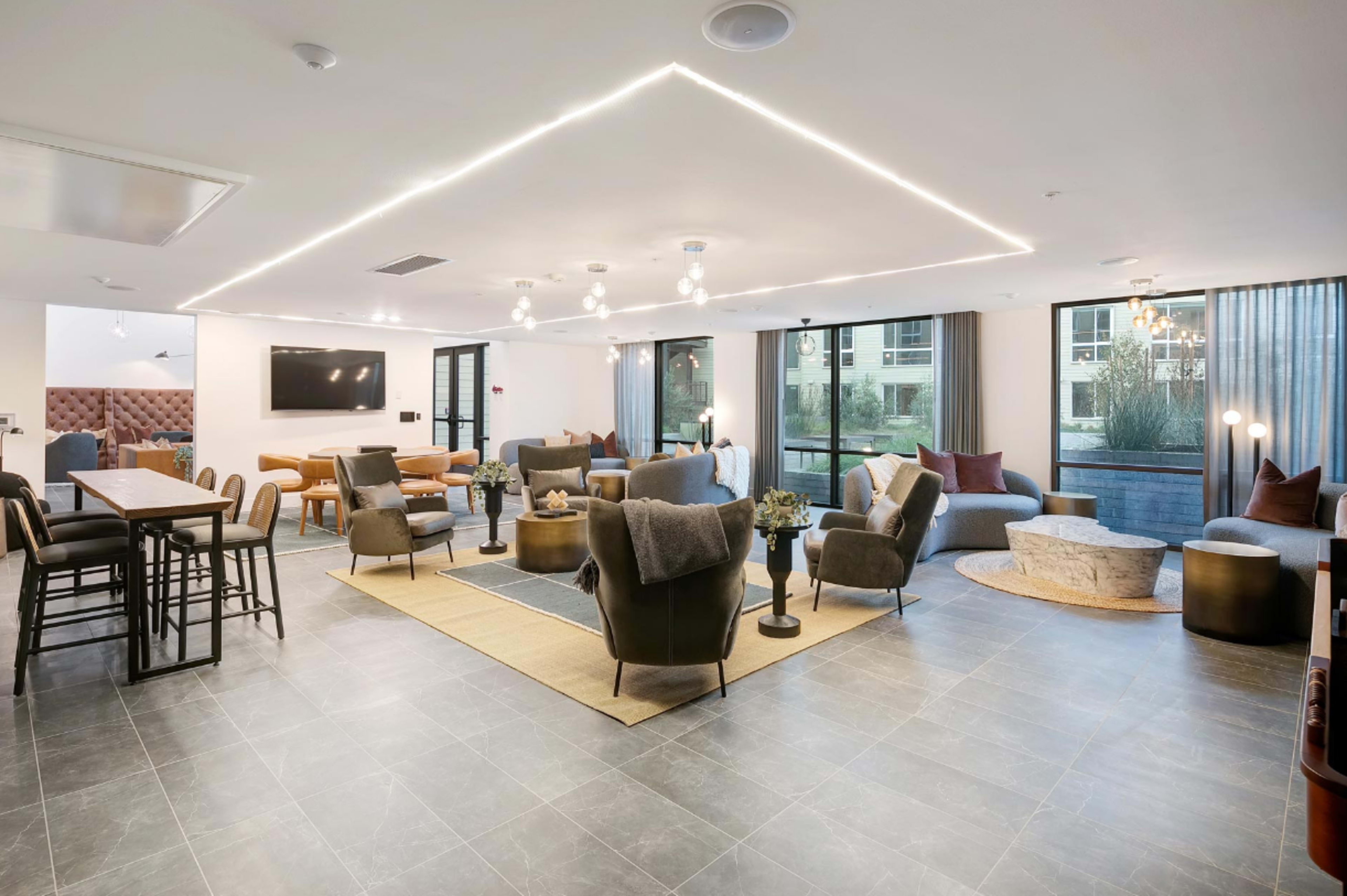 A modern common area with various seating arrangements, including armchairs and a dining table, illuminated by recessed lighting and large windows.