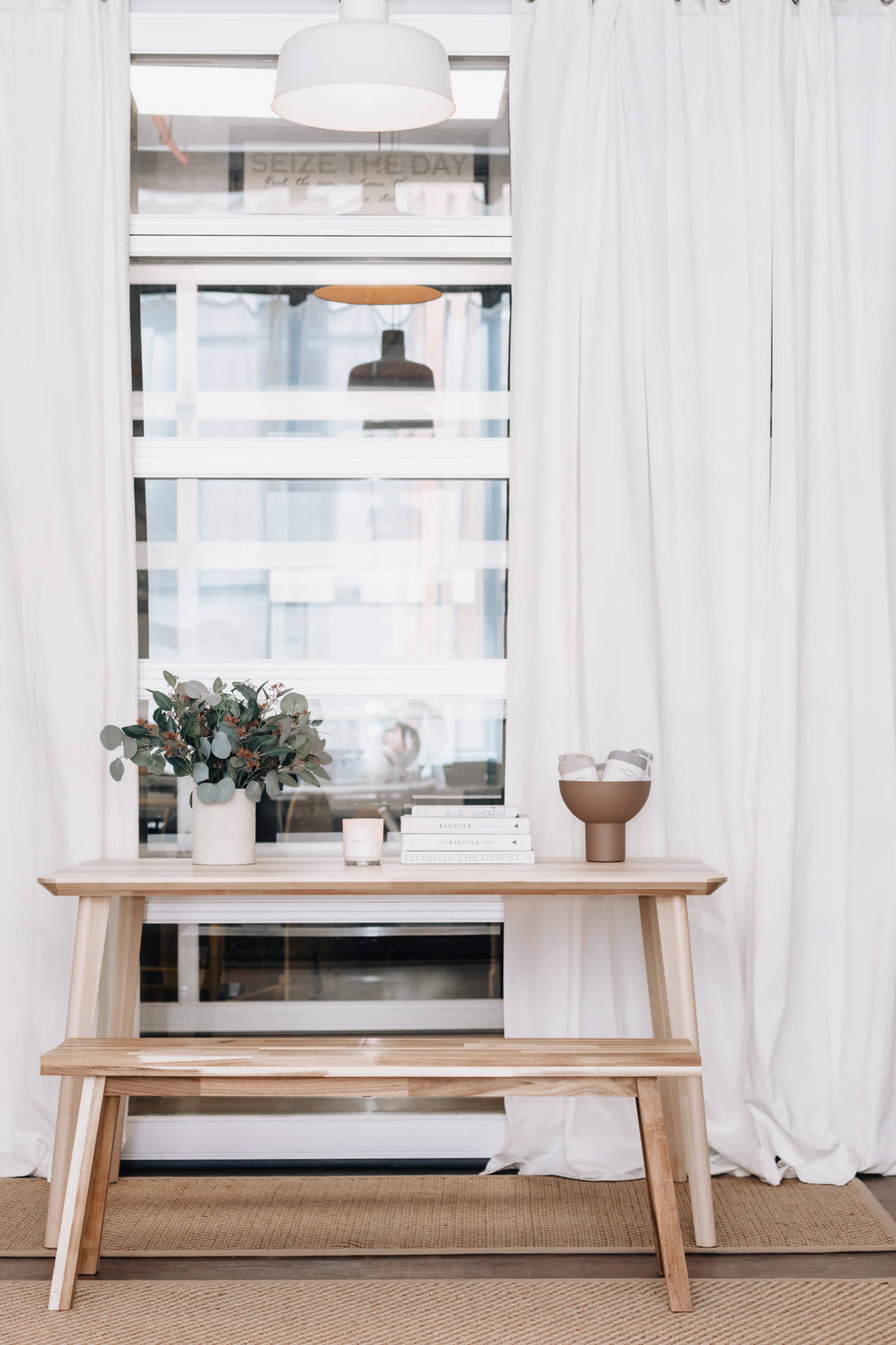 A wooden table with a vase of flowers and a bowl sits in front of sheer white curtains and a glass wall.