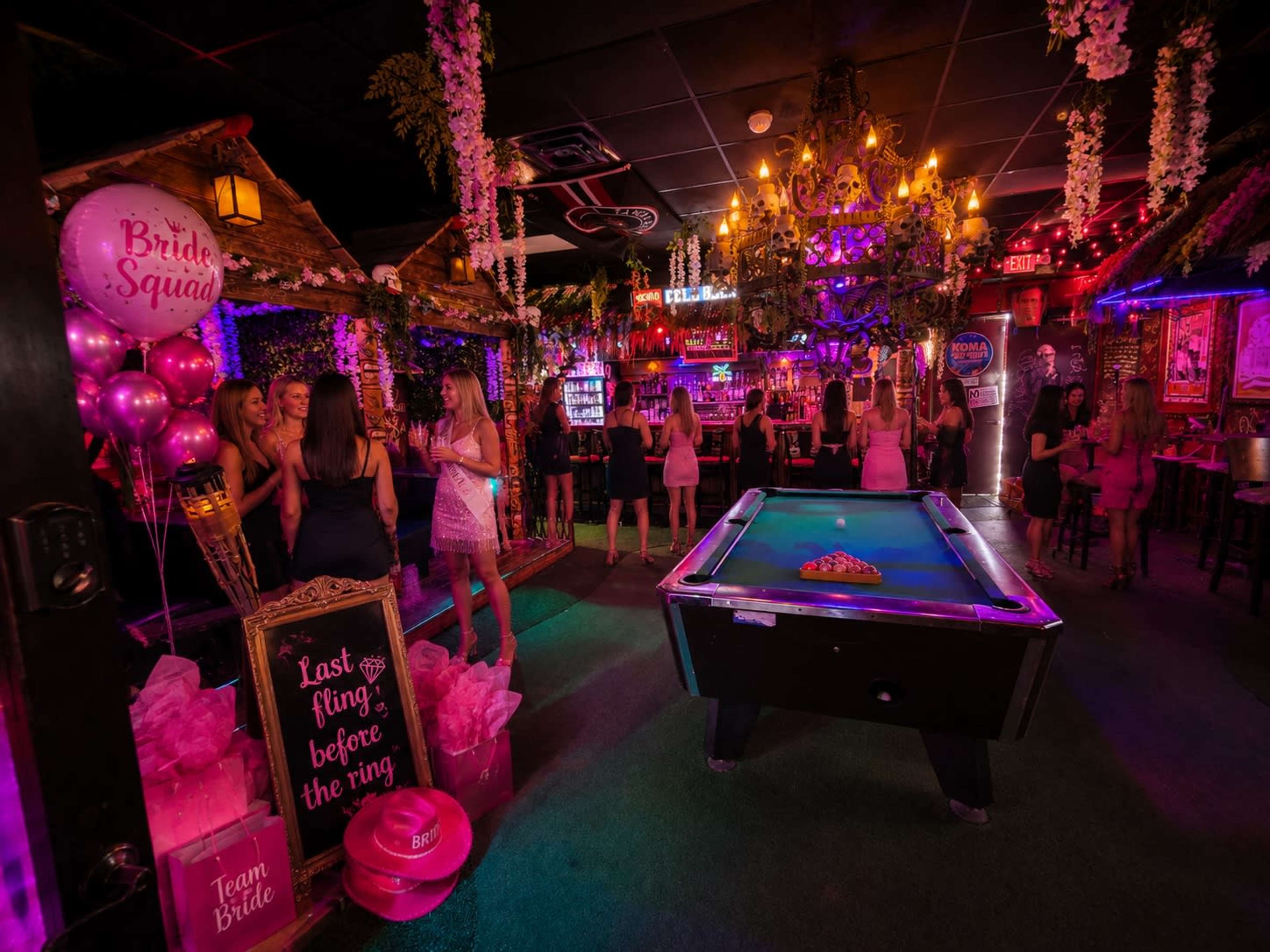 A lively indoor venue is decorated with balloons and flowers, where a group of women celebrate a bridal party around a pool table and bar.