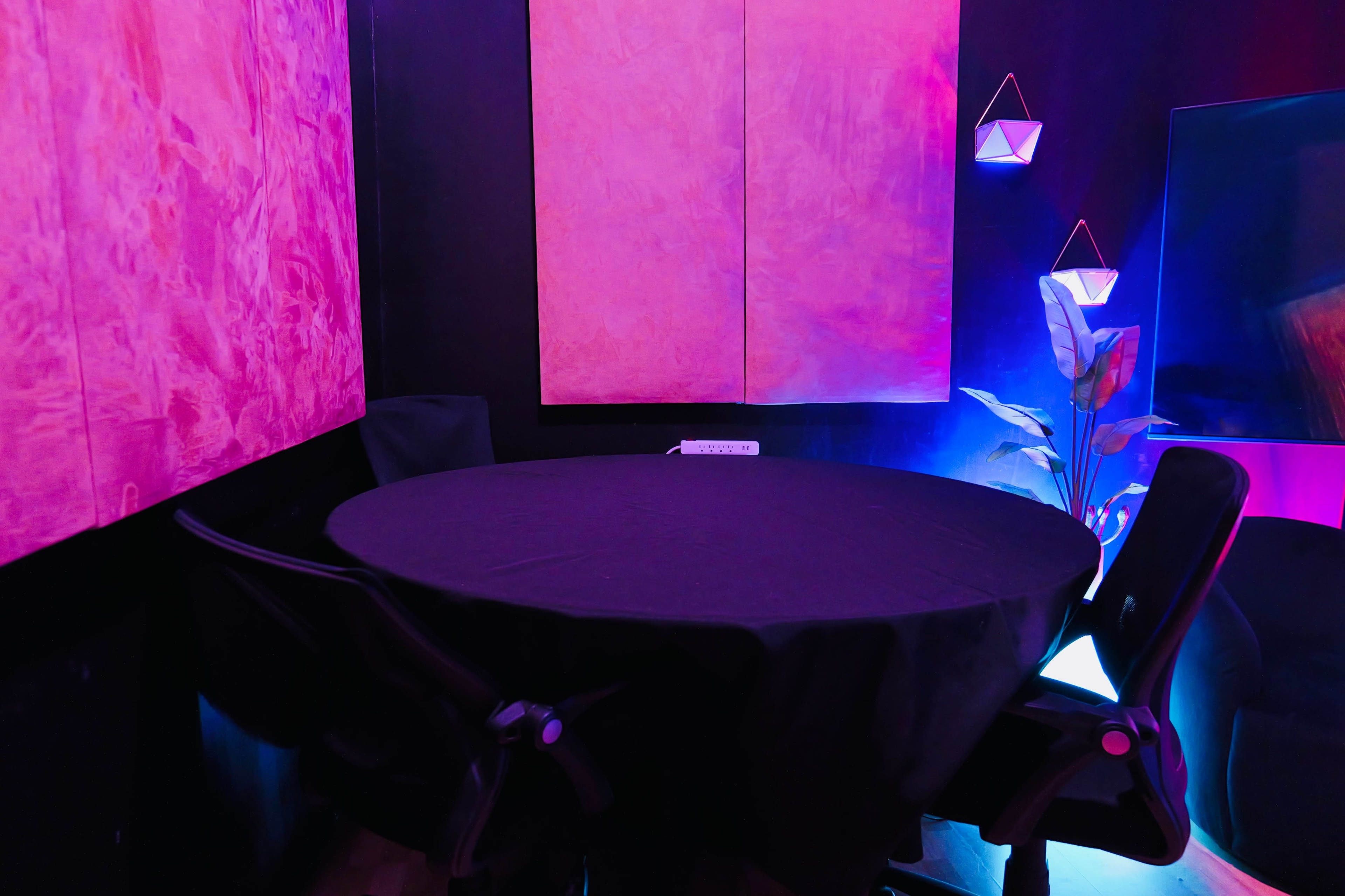 A round black table is set against walls featuring pink artwork, with two black chairs and a small plant illuminated by colored lights.