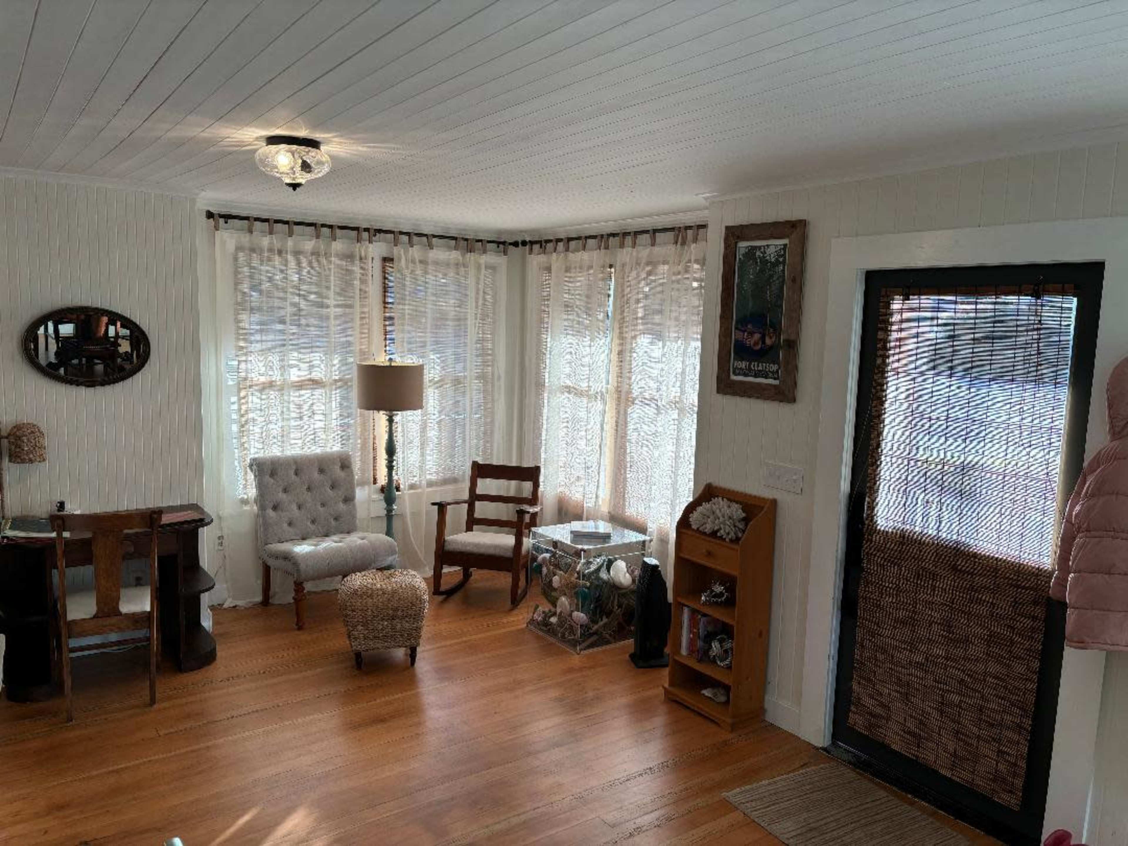 The image shows a sunlit room with wooden flooring, featuring a chair, a small table, a lamp, and a large window dressed with sheer curtains.
