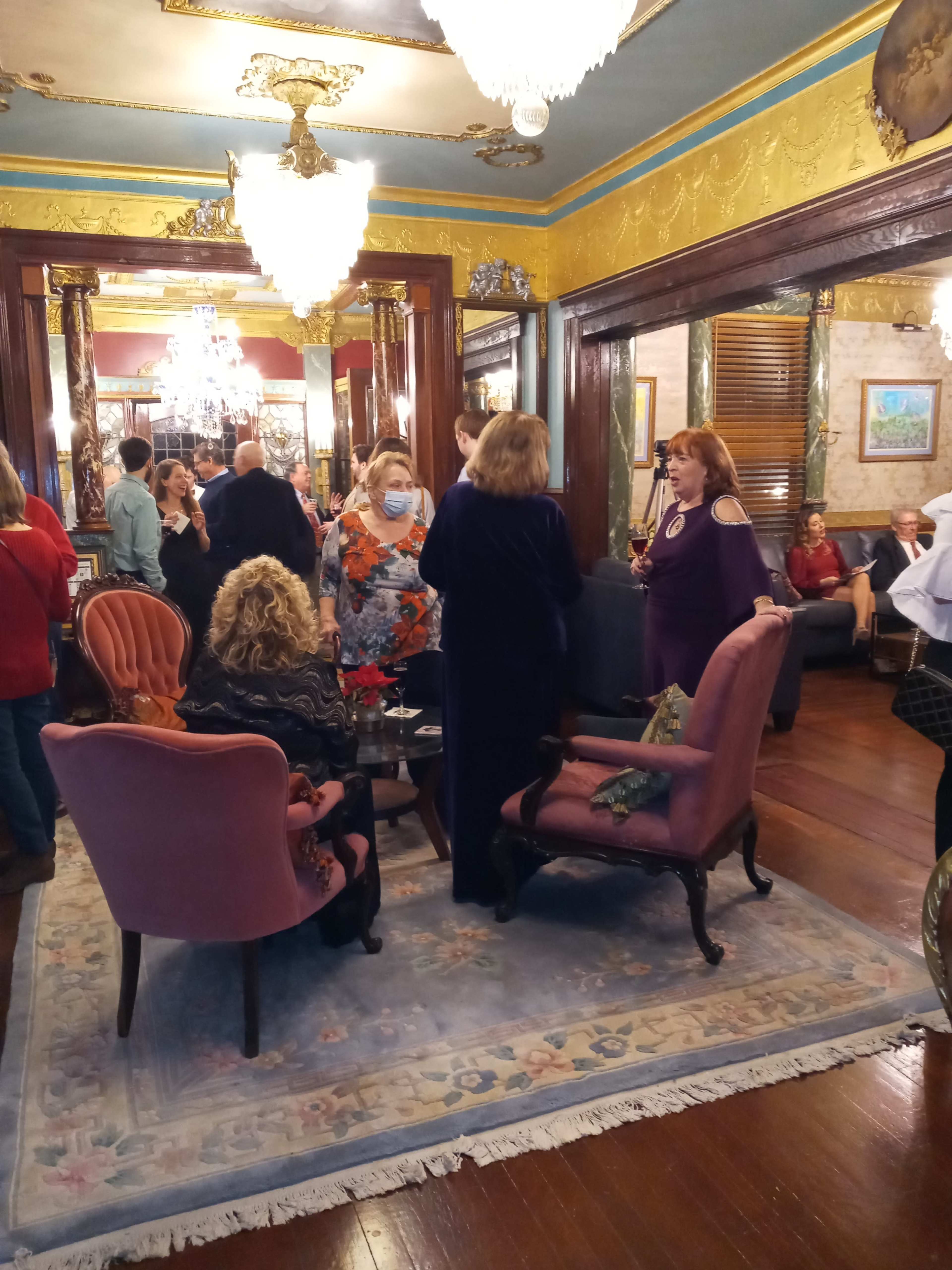 A group of people mingles in a warmly lit, elegantly decorated room with vintage furniture and ornate chandeliers.