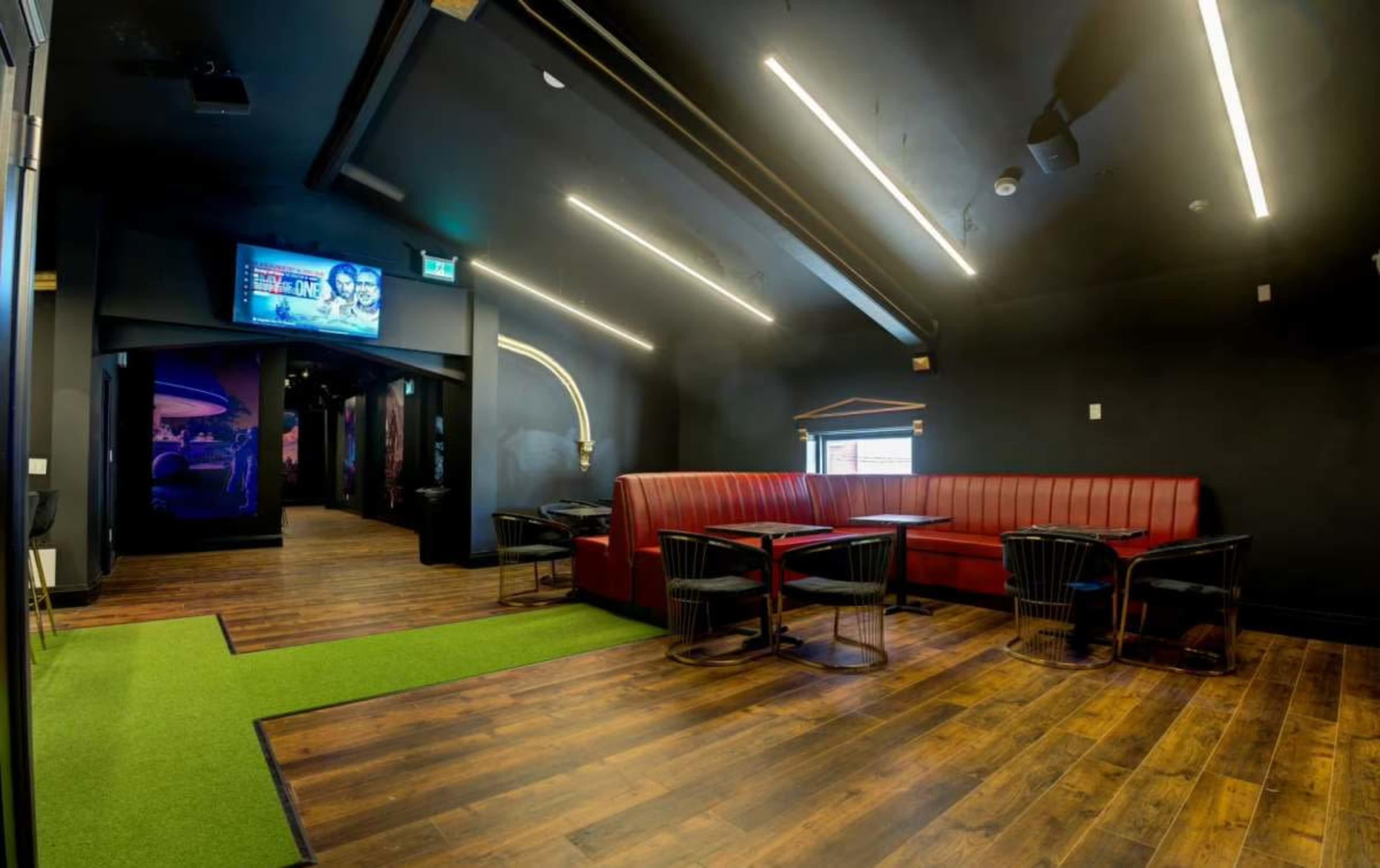 The image shows a modern interior of a lounge with a red upholstered seating area, several black tables and chairs, and a green carpet leading to a hallway.