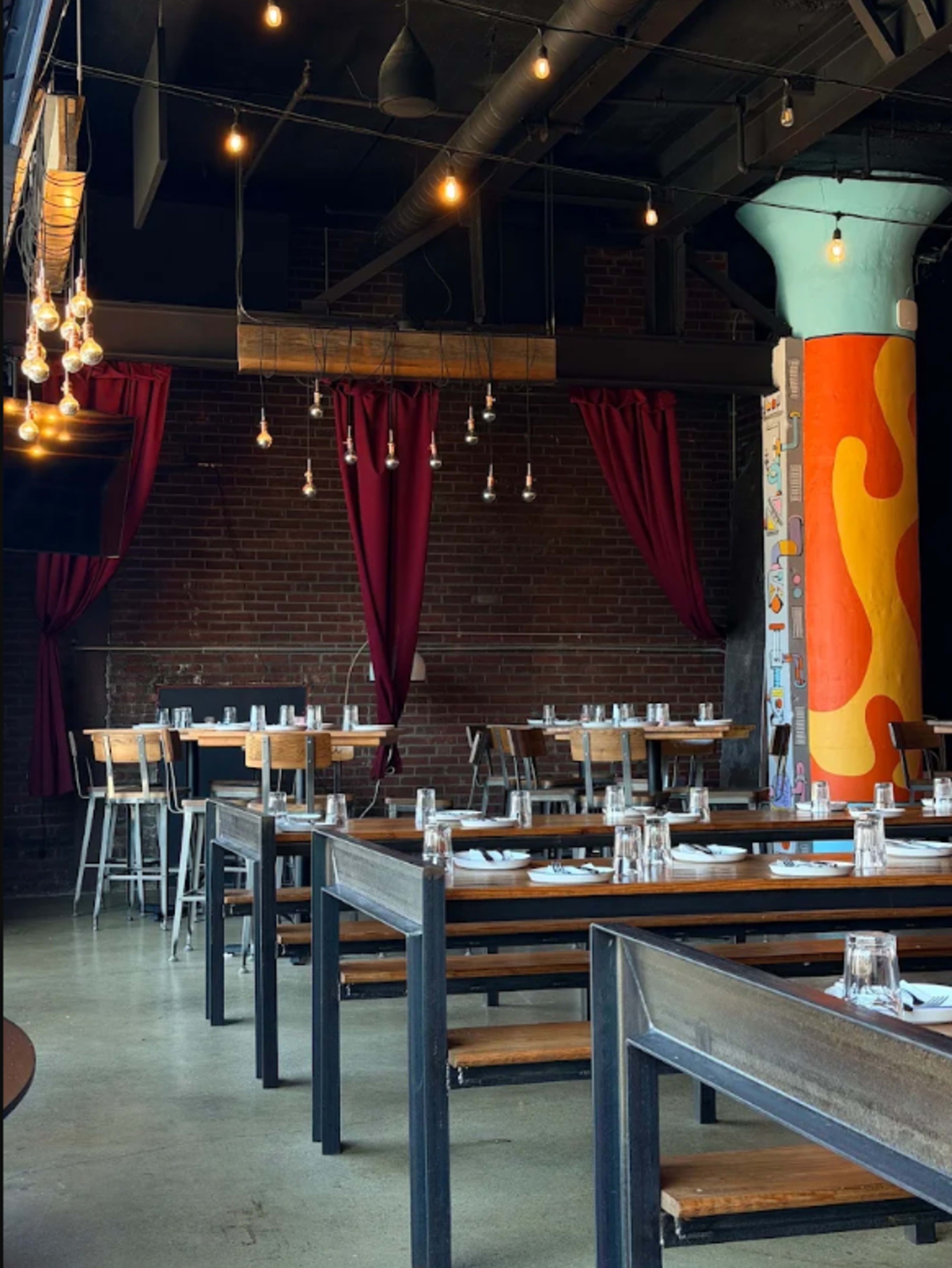 The interior of a restaurant features wooden tables with metal chairs, exposed brick walls, and colorful decorative columns, illuminated by hanging light bulbs.