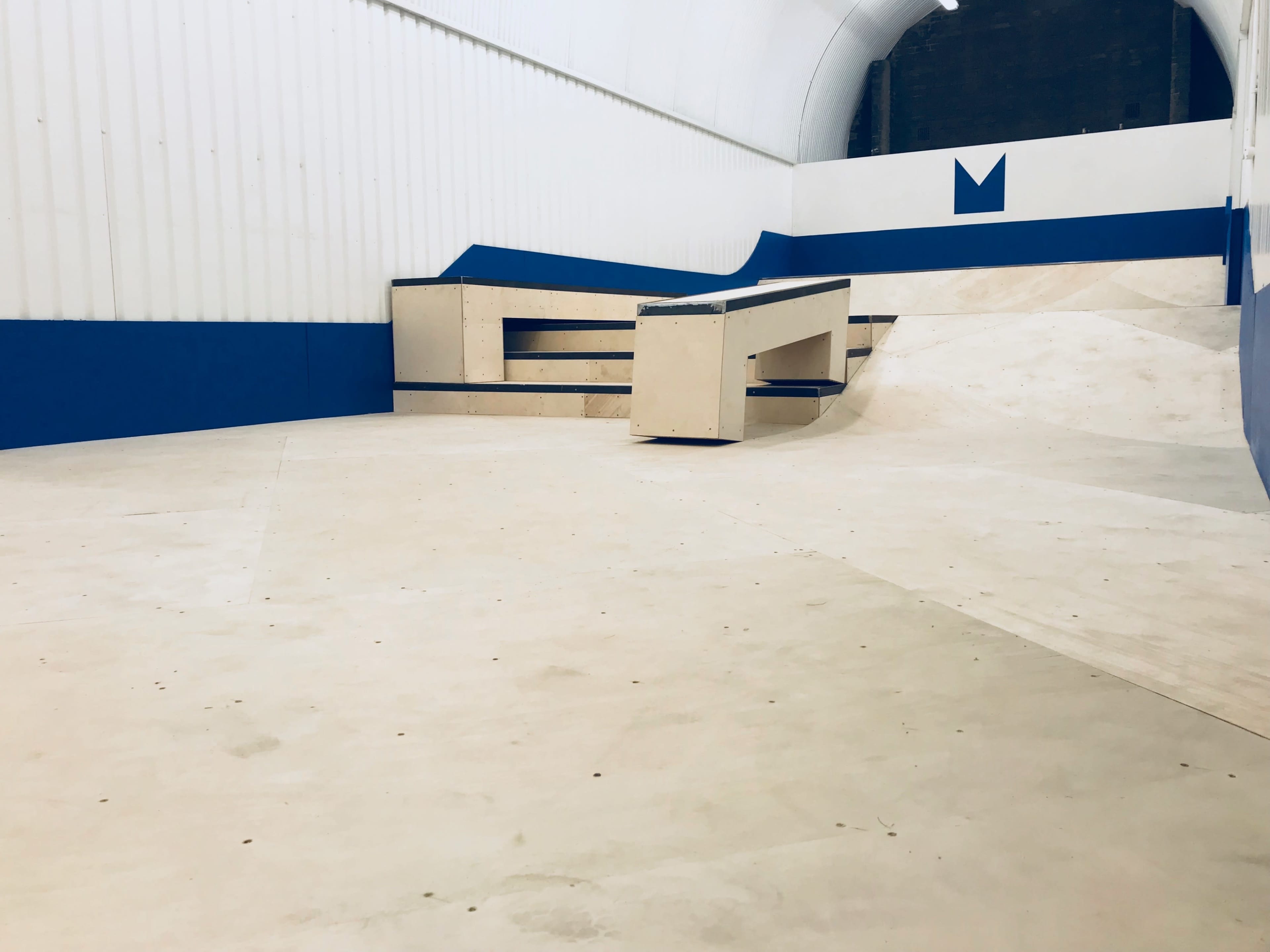 A skatepark with a smooth, light-colored floor, ramps, and a wooden obstacle surrounded by a blue and white wall.