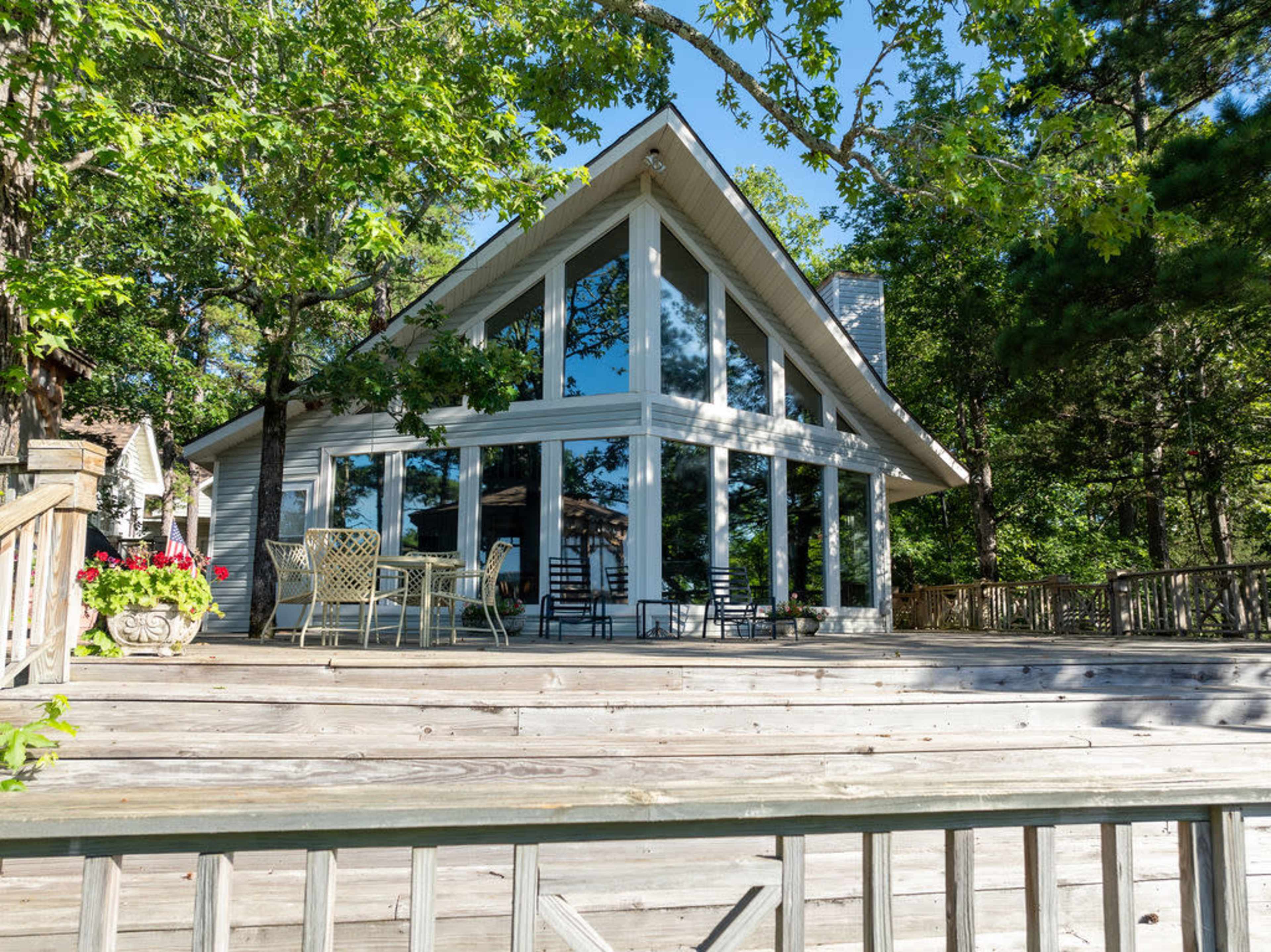 A modern wooden house with large glass windows is surrounded by trees and features a spacious deck with outdoor furniture.