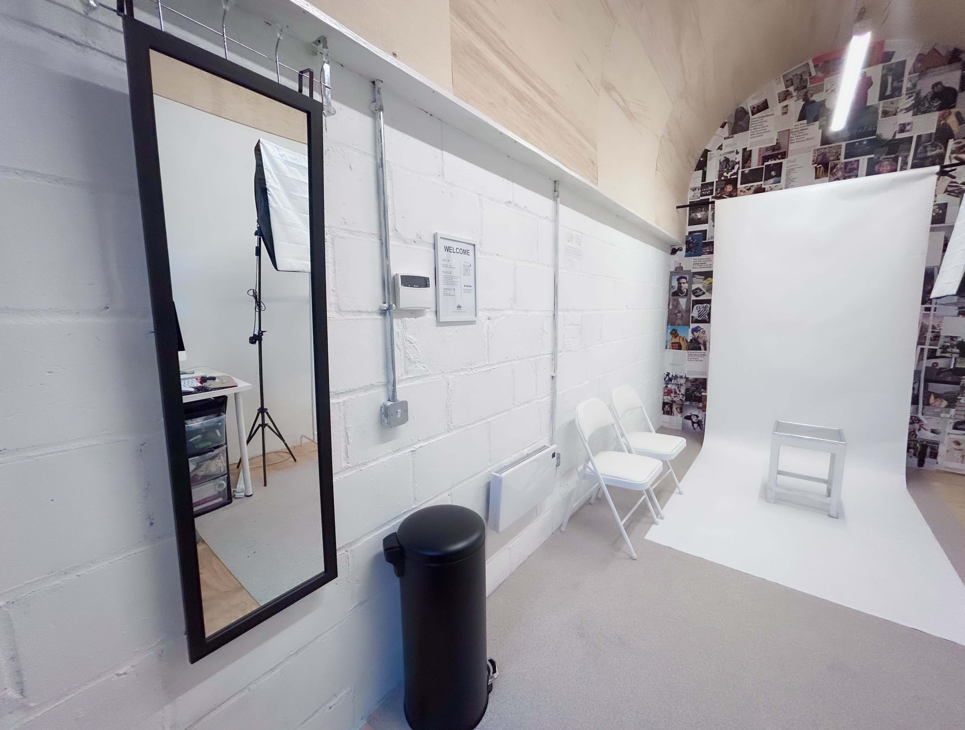 The image shows a photo studio with a large mirror, a white backdrop, a chair, and a trash can against a wall covered in black-and-white photos.