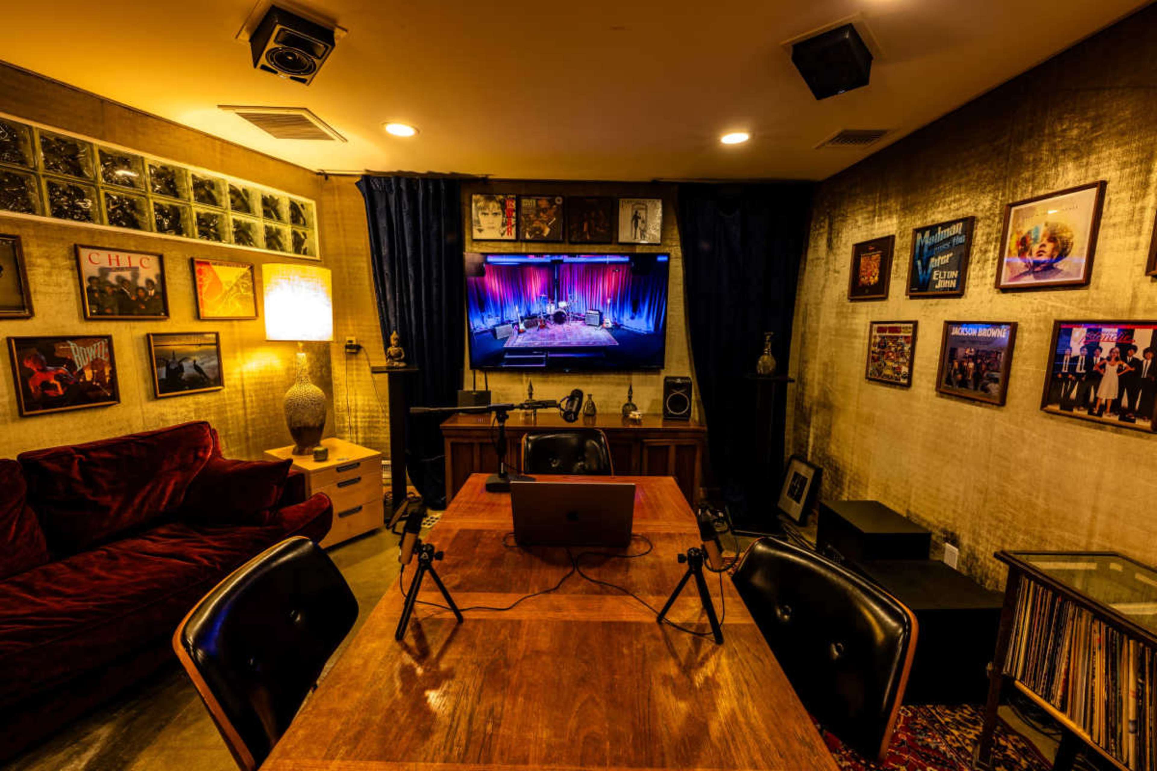 The image shows a dimly lit room with a large screen displaying a performance, surrounded by framed records and a wooden dining table with microphones set up.