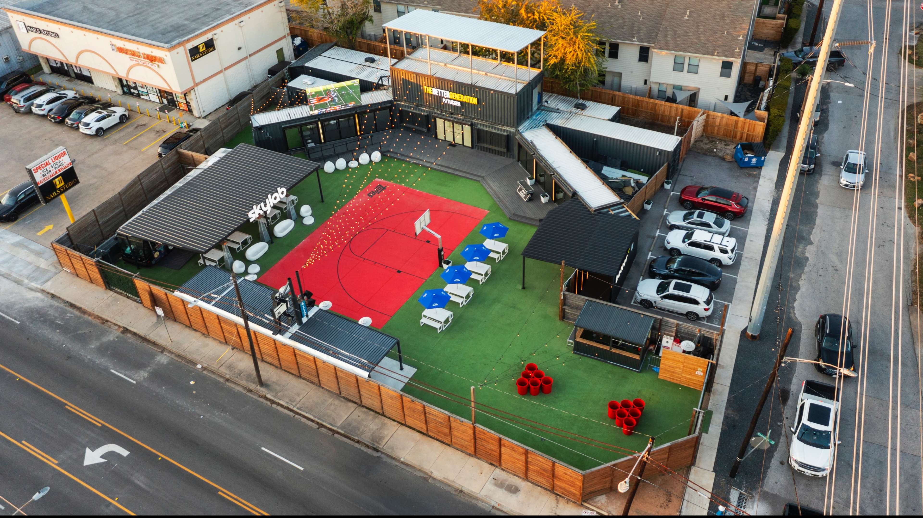 The image shows an outdoor recreational area with a red and green turf layout, surrounded by seating arrangements and parked cars nearby.