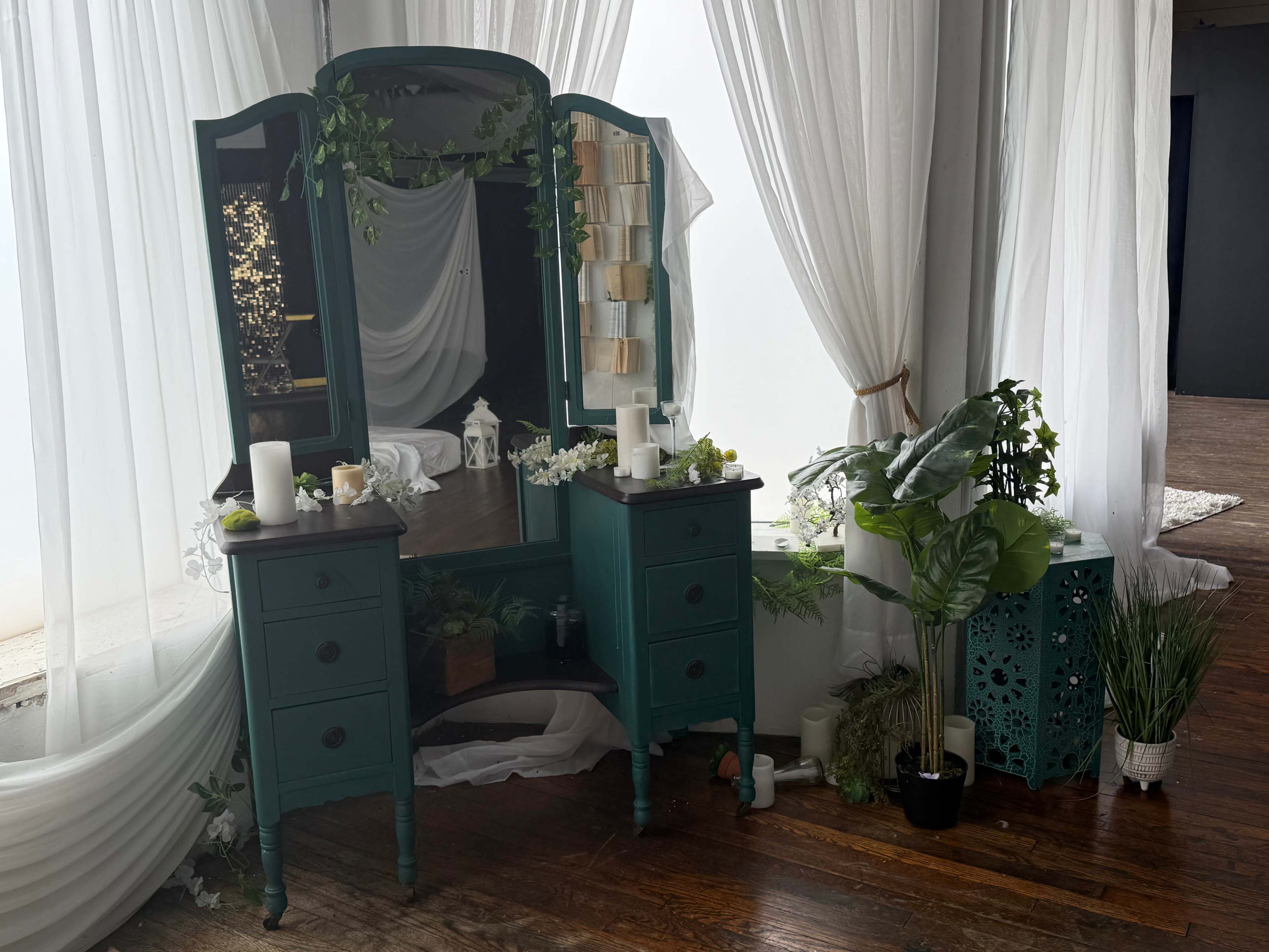 The image shows a teal vanity table with a large mirror, flanked by potted plants and decorative elements, set against sheer white curtains in a bright, airy room.