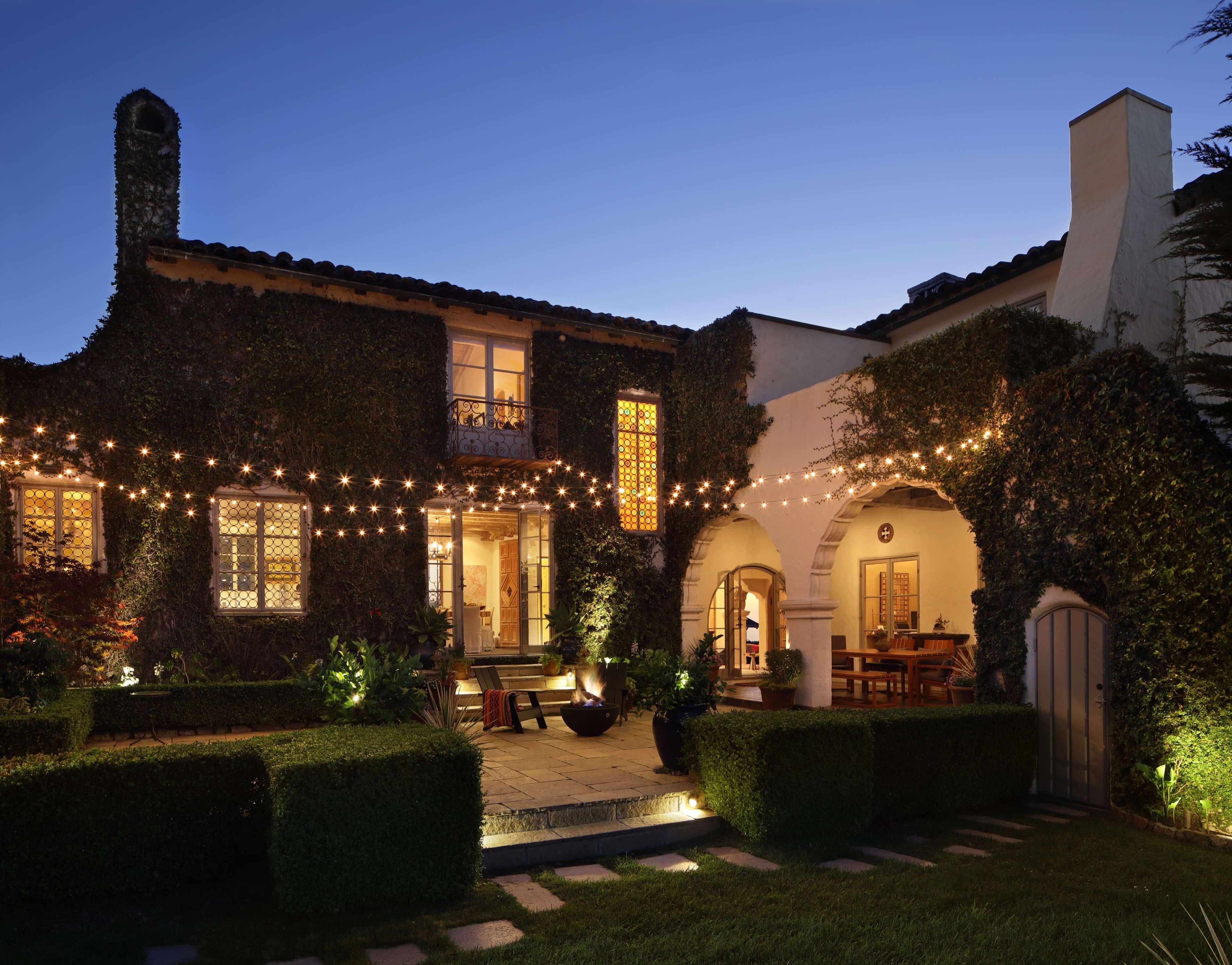 A two-story house with ivy-covered walls is illuminated by string lights in the backyard at dusk.