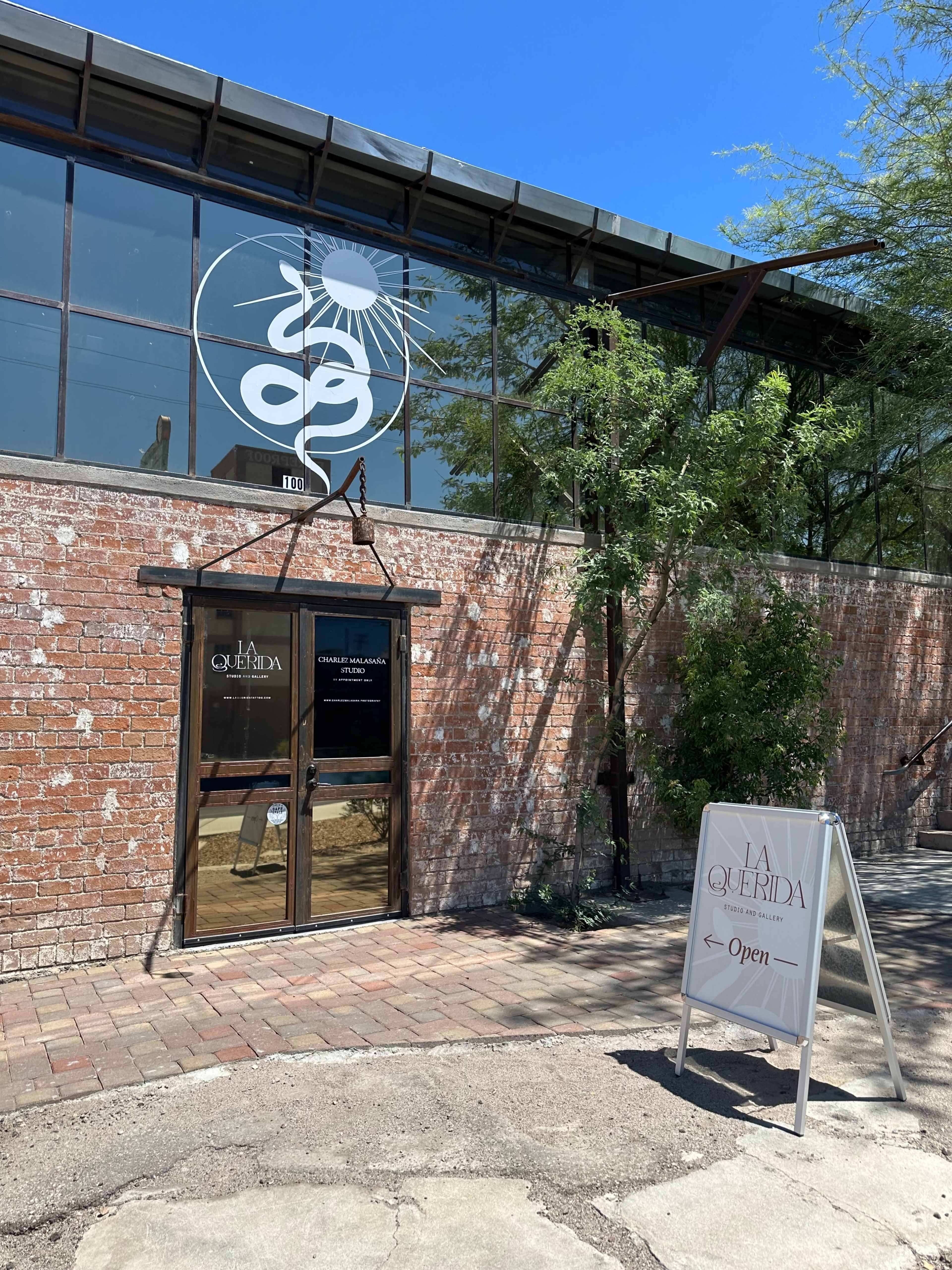 A brick building with large glass windows features a sign for "La Querida" and an open status sign.