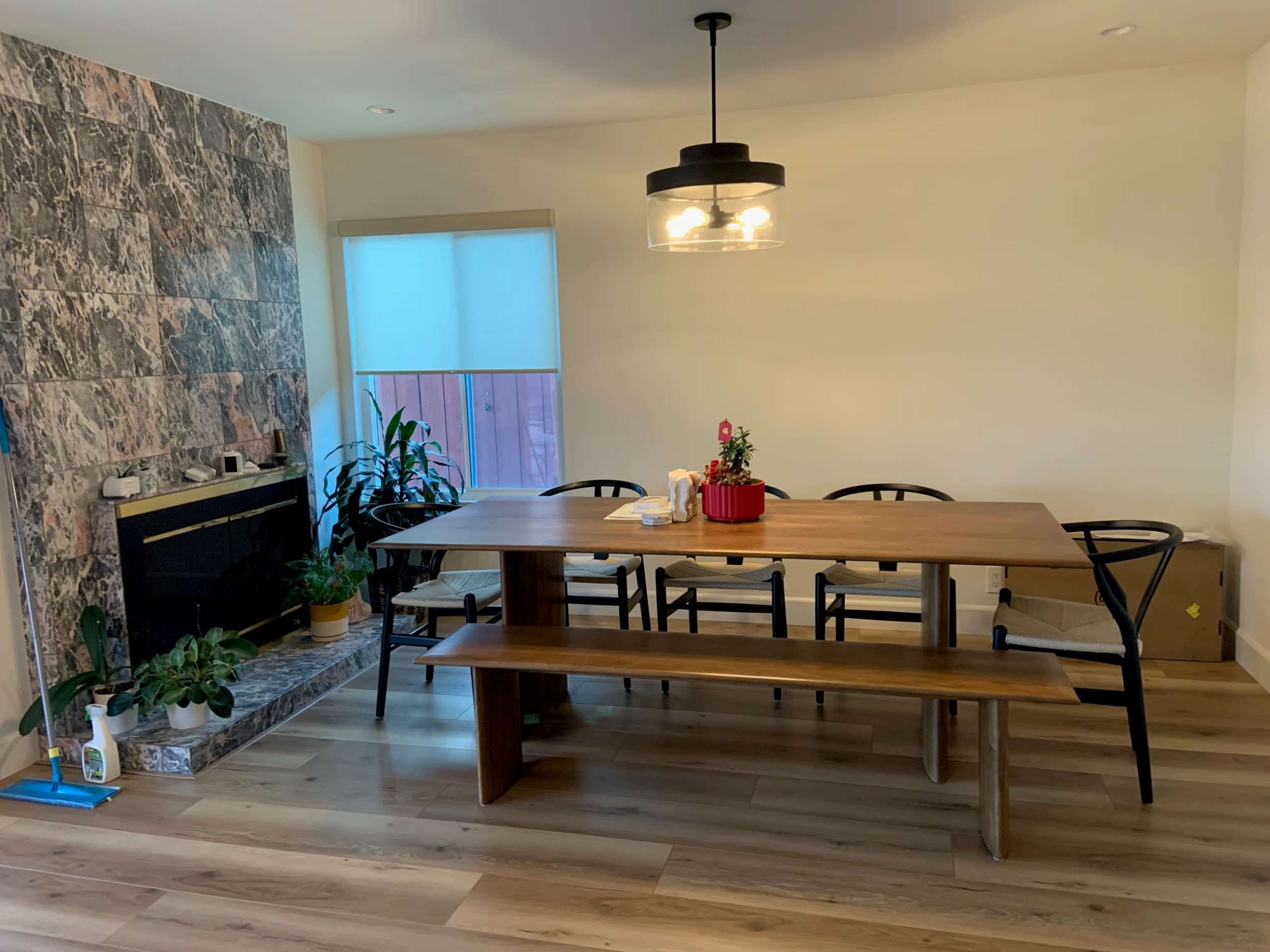 A spacious dining area features a long wooden table with black chairs, a modern light fixture, and a stone-patterned wall beside a fireplace, adorned with potted plants.