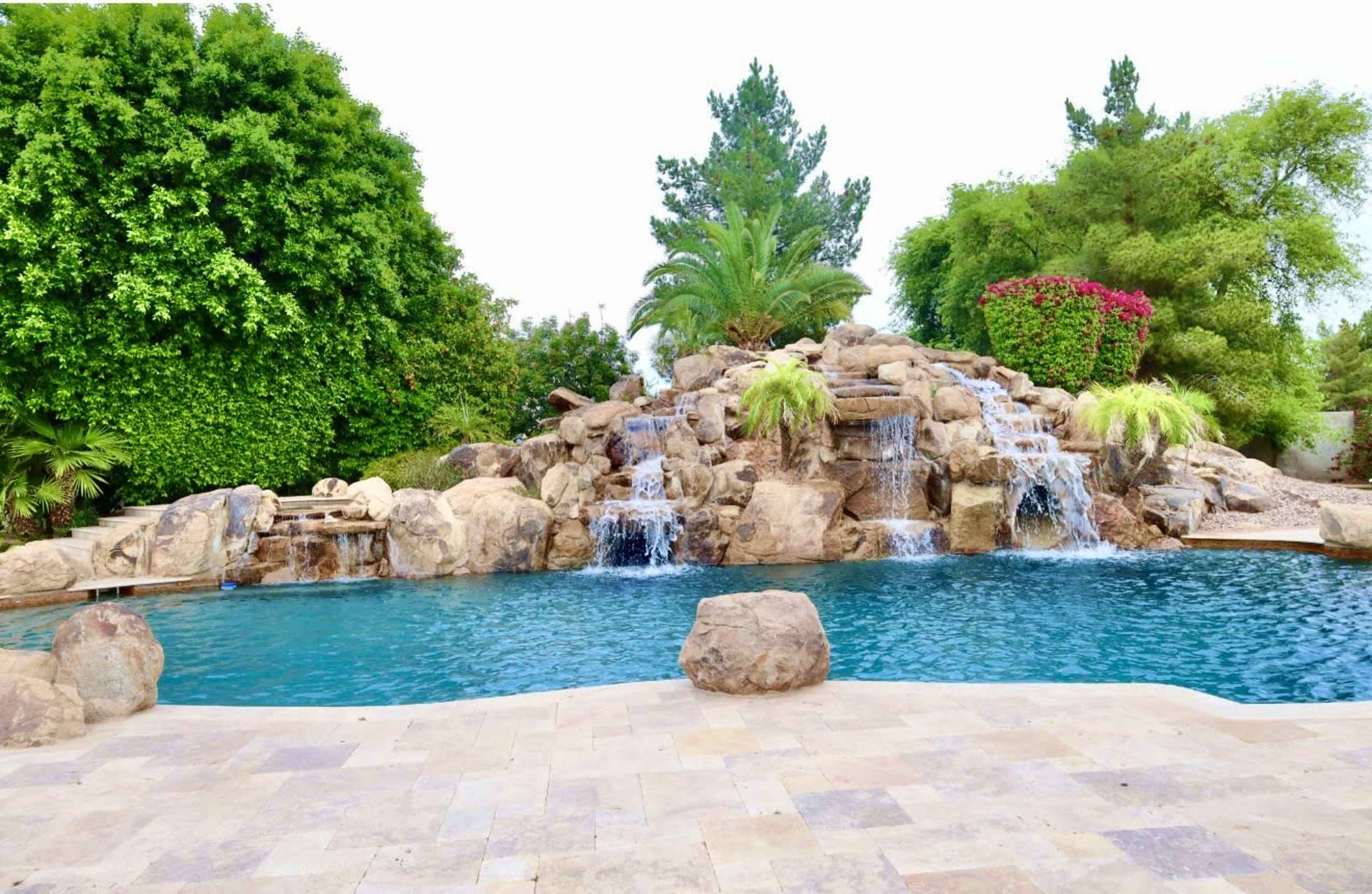 The image shows a swimming pool with a rock waterfall surrounded by lush greenery and colorful plants.