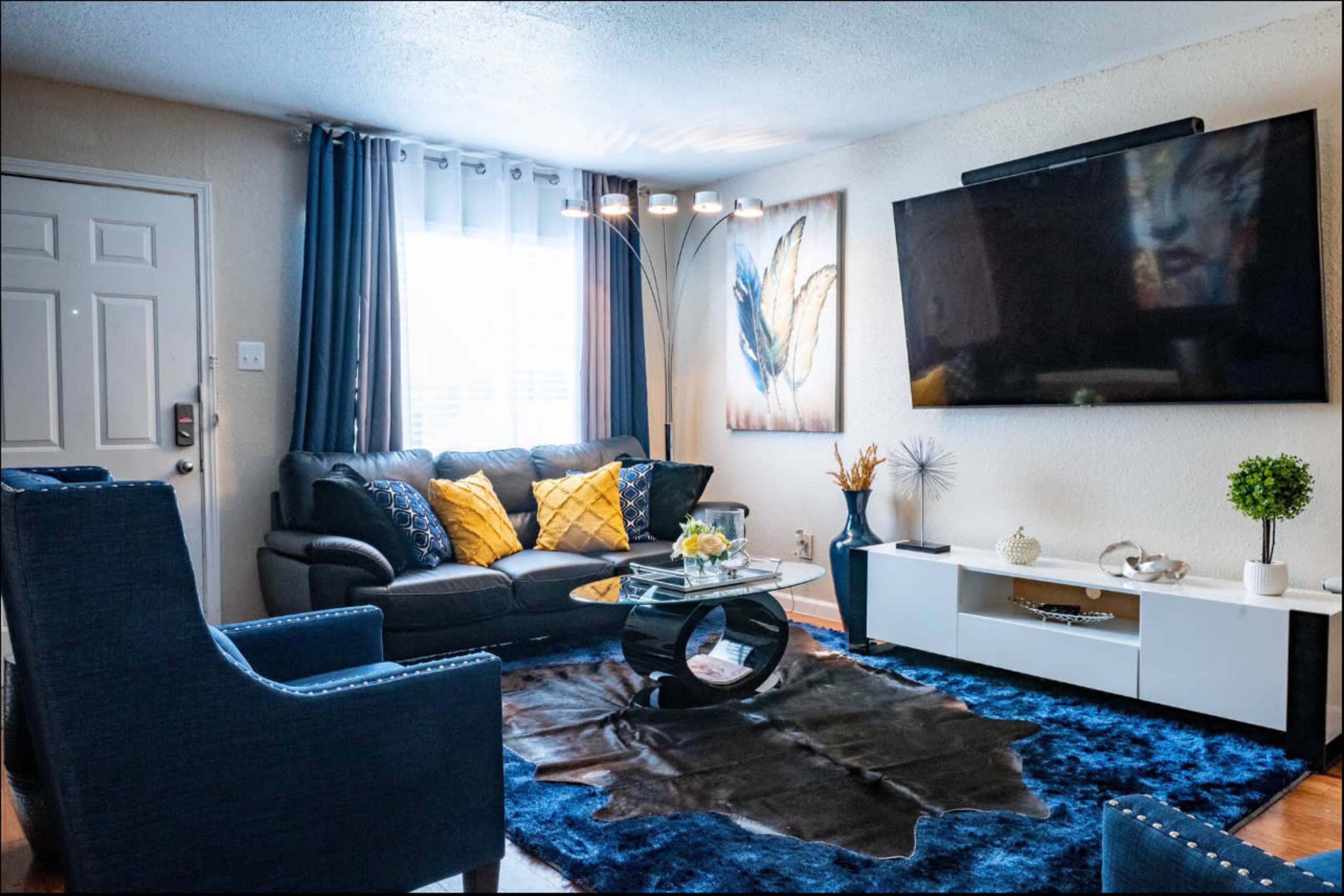 The image shows a living room with a black sofa, yellow and blue decorative pillows, a glass coffee table on a blue rug, and a large wall-mounted television.