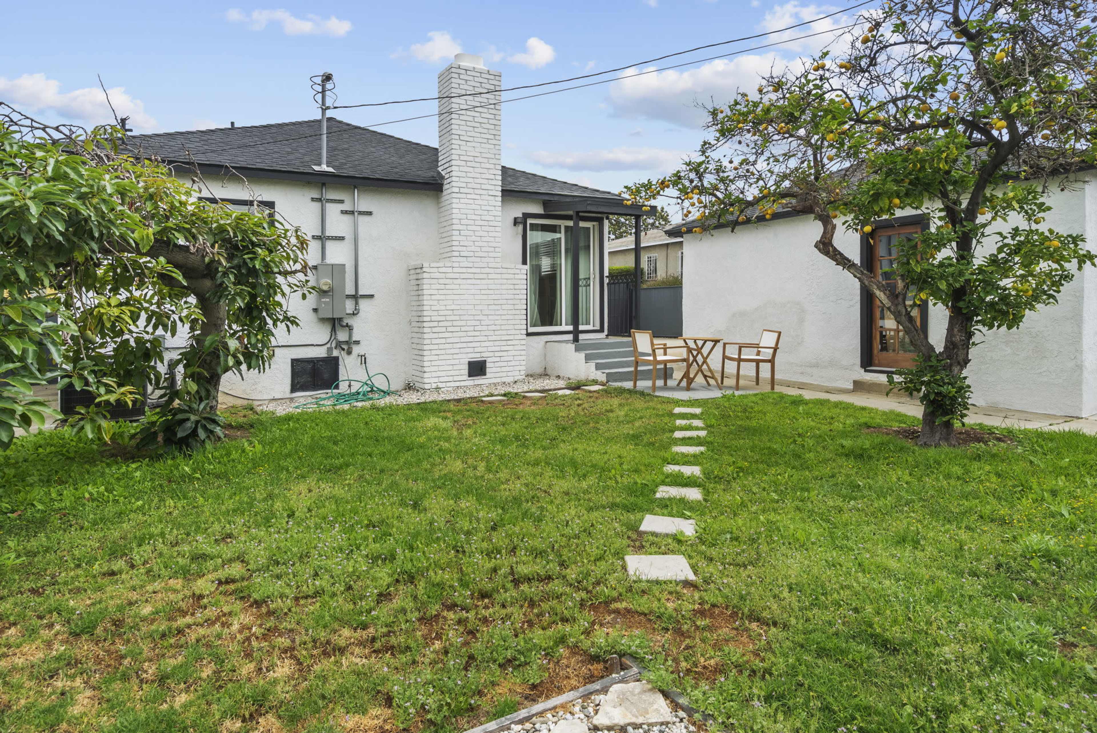 The image shows a backyard with a grassy area, a stone pathway leading towards a small patio with outdoor furniture, and trees on either side.
