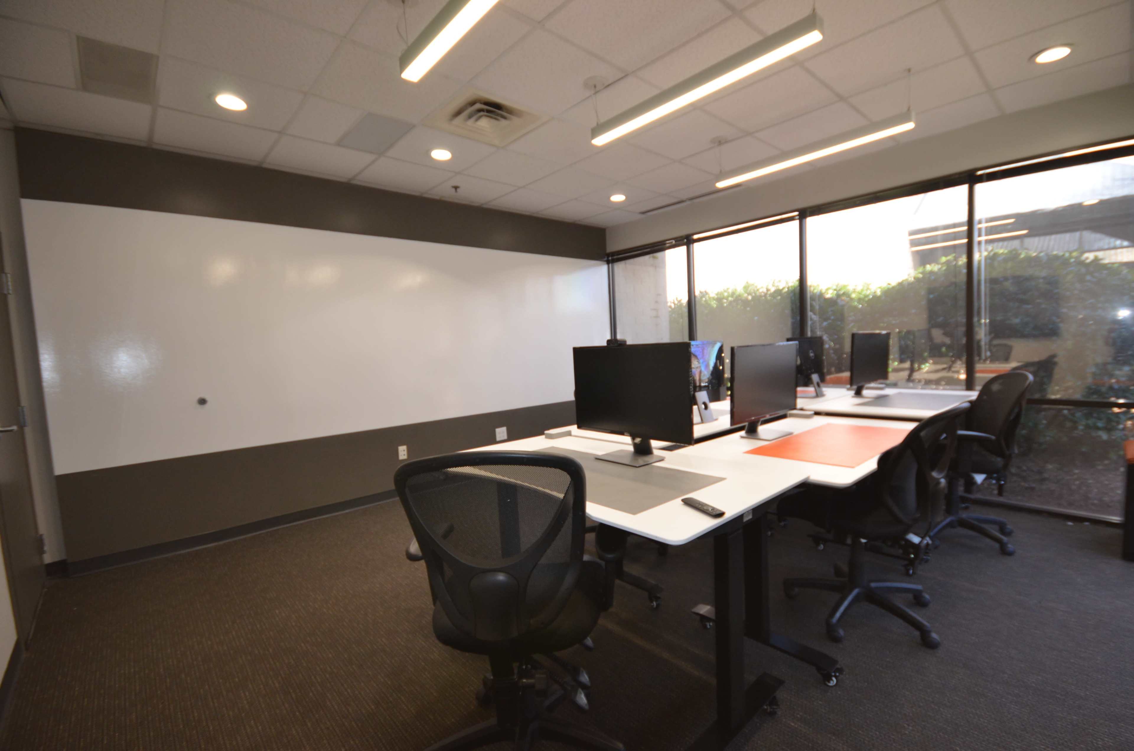 A modern office space features four computer stations facing a large window with outdoor greenery and a whiteboard on the wall.