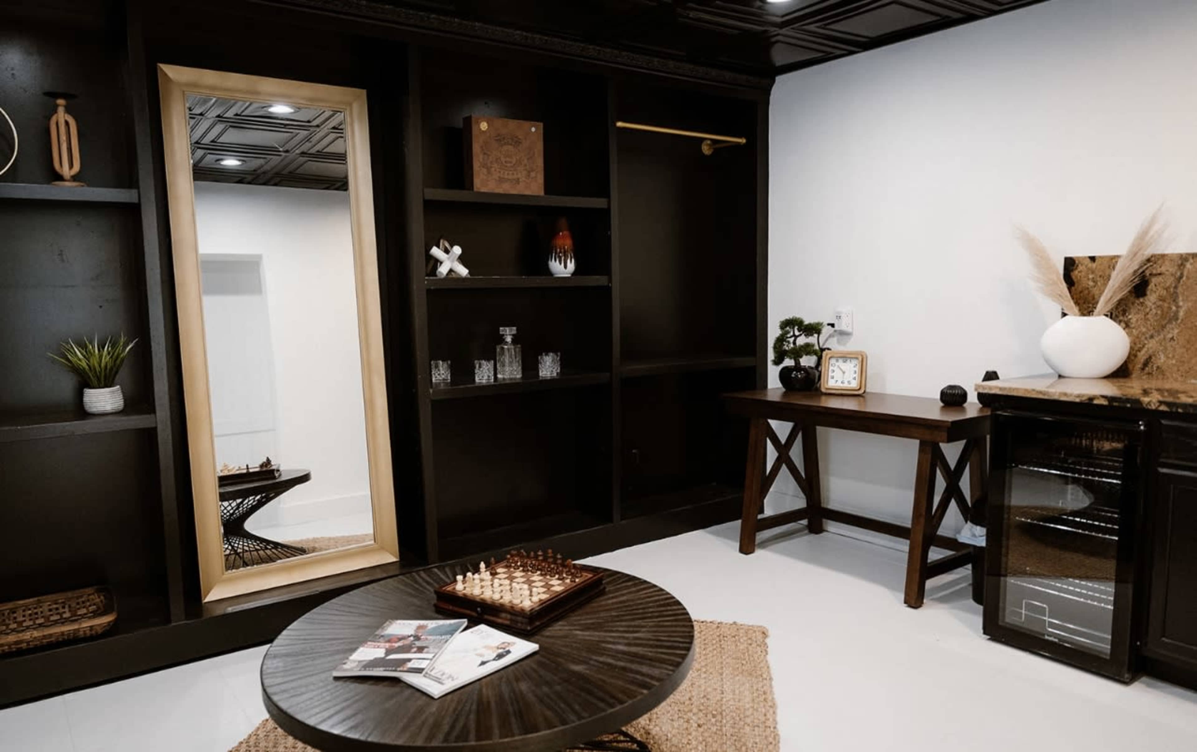 The image shows a modern room featuring black shelving, a decorative mirror, a wooden table with a clock, and a small chess set on a round coffee table.