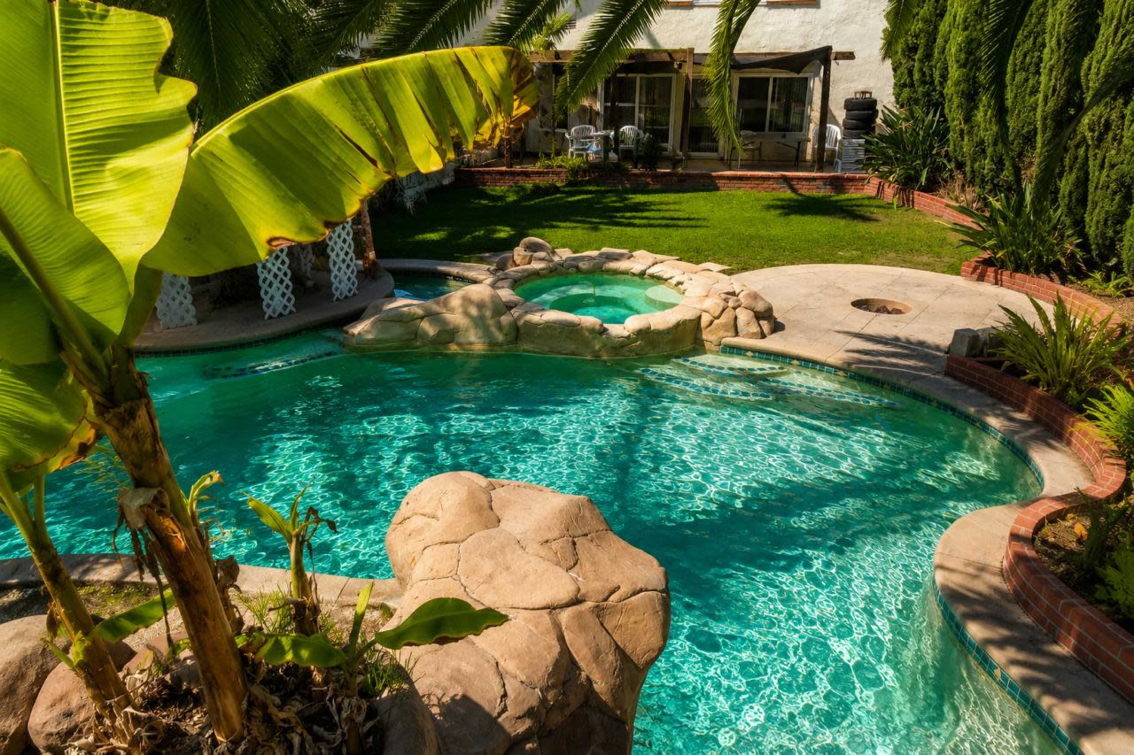 The image shows a swimming pool with a rocky spa area surrounded by tropical plants and a well-kept lawn.