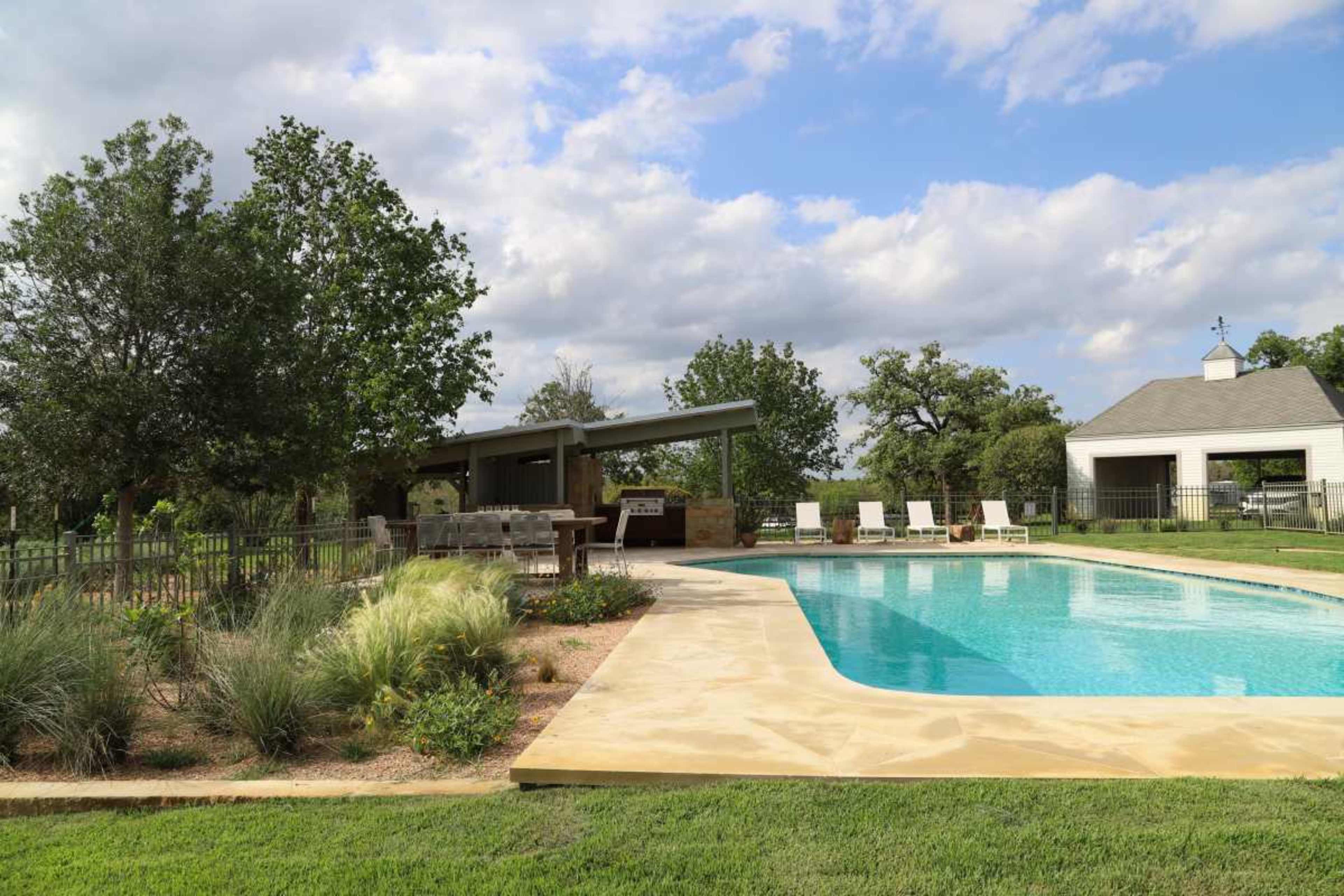 A clean swimming pool surrounded by landscaped greenery and a shaded seating area in a residential backyard.