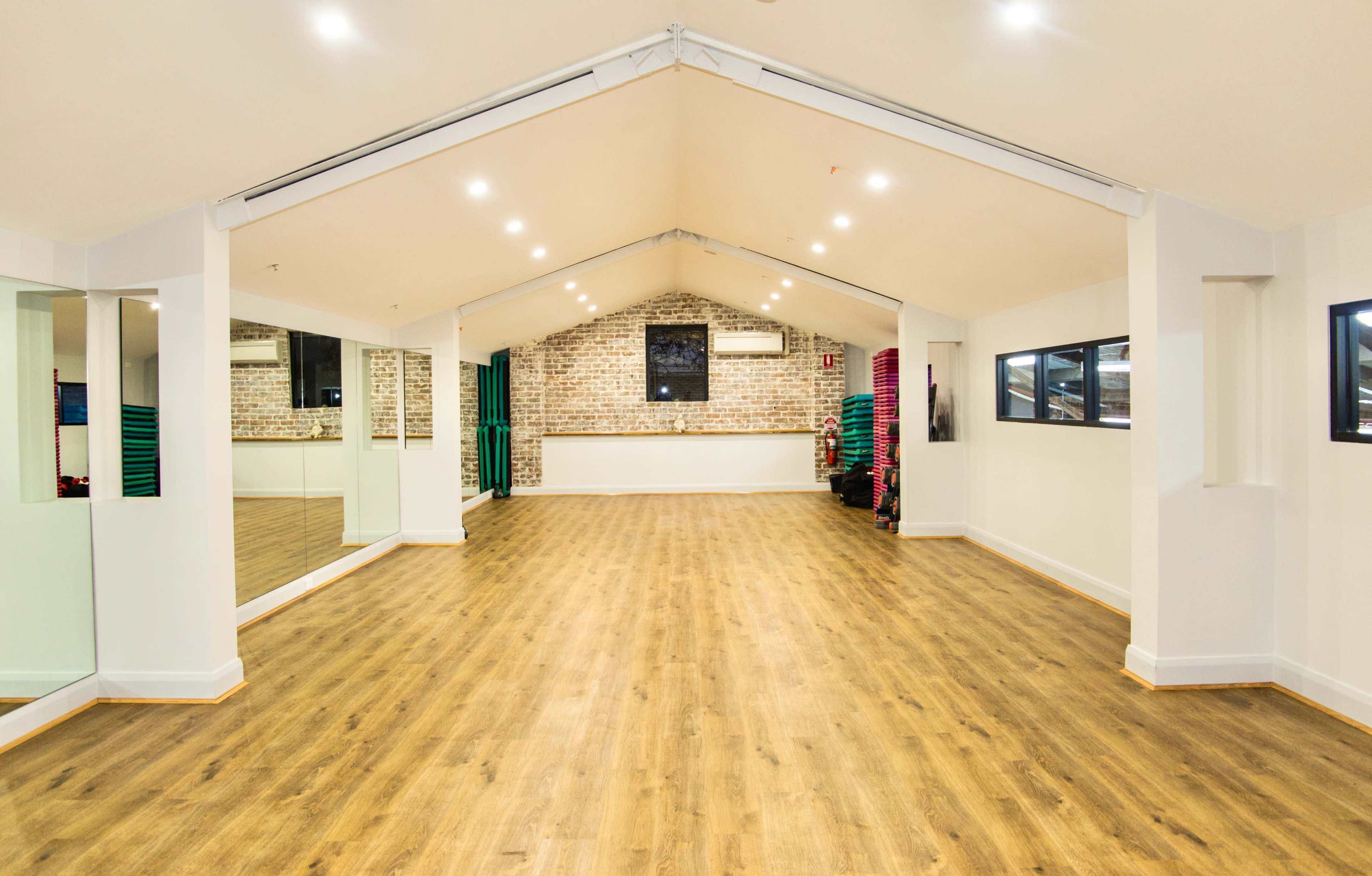 A spacious fitness studio with wooden floors, white walls, and ample natural light from large windows.