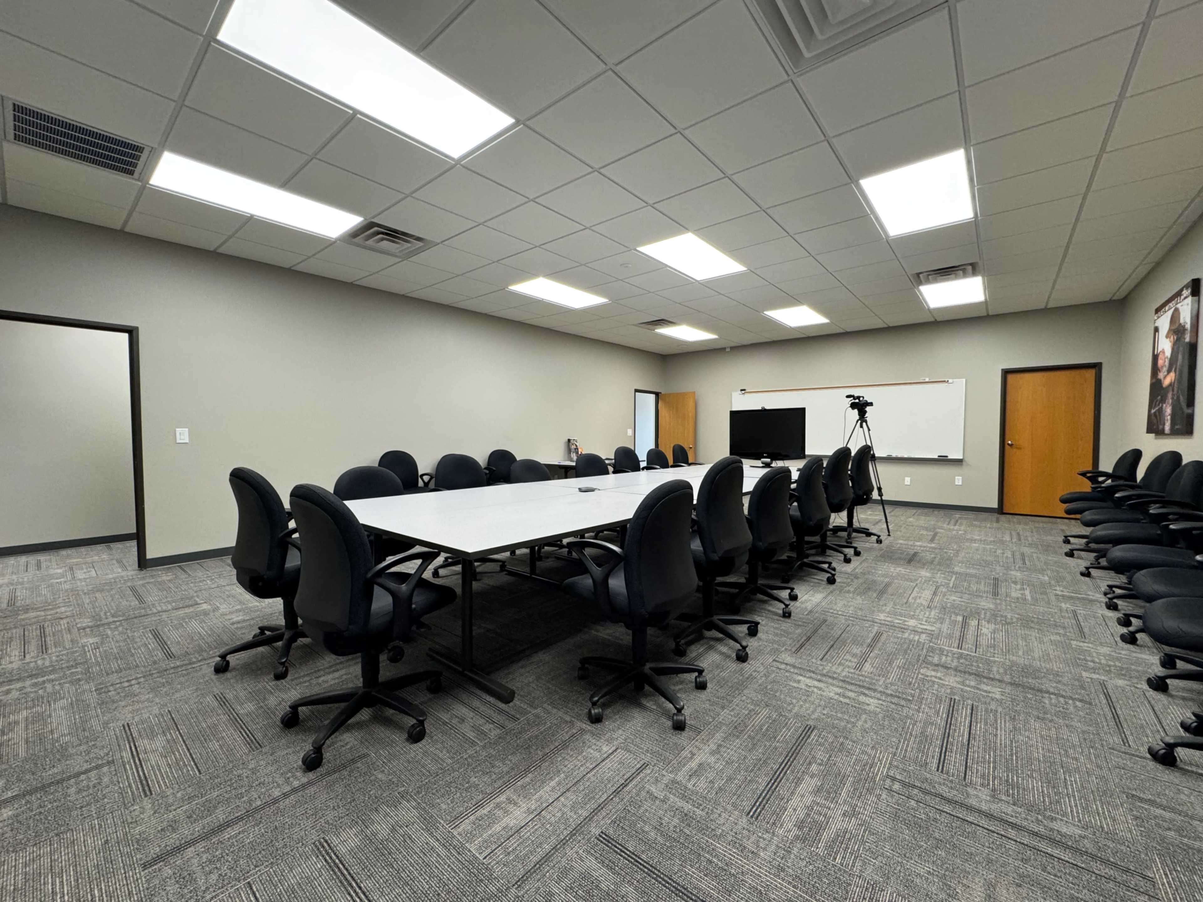 A spacious conference room features a long rectangular table surrounded by black rolling chairs, with additional chairs aligned against the walls.