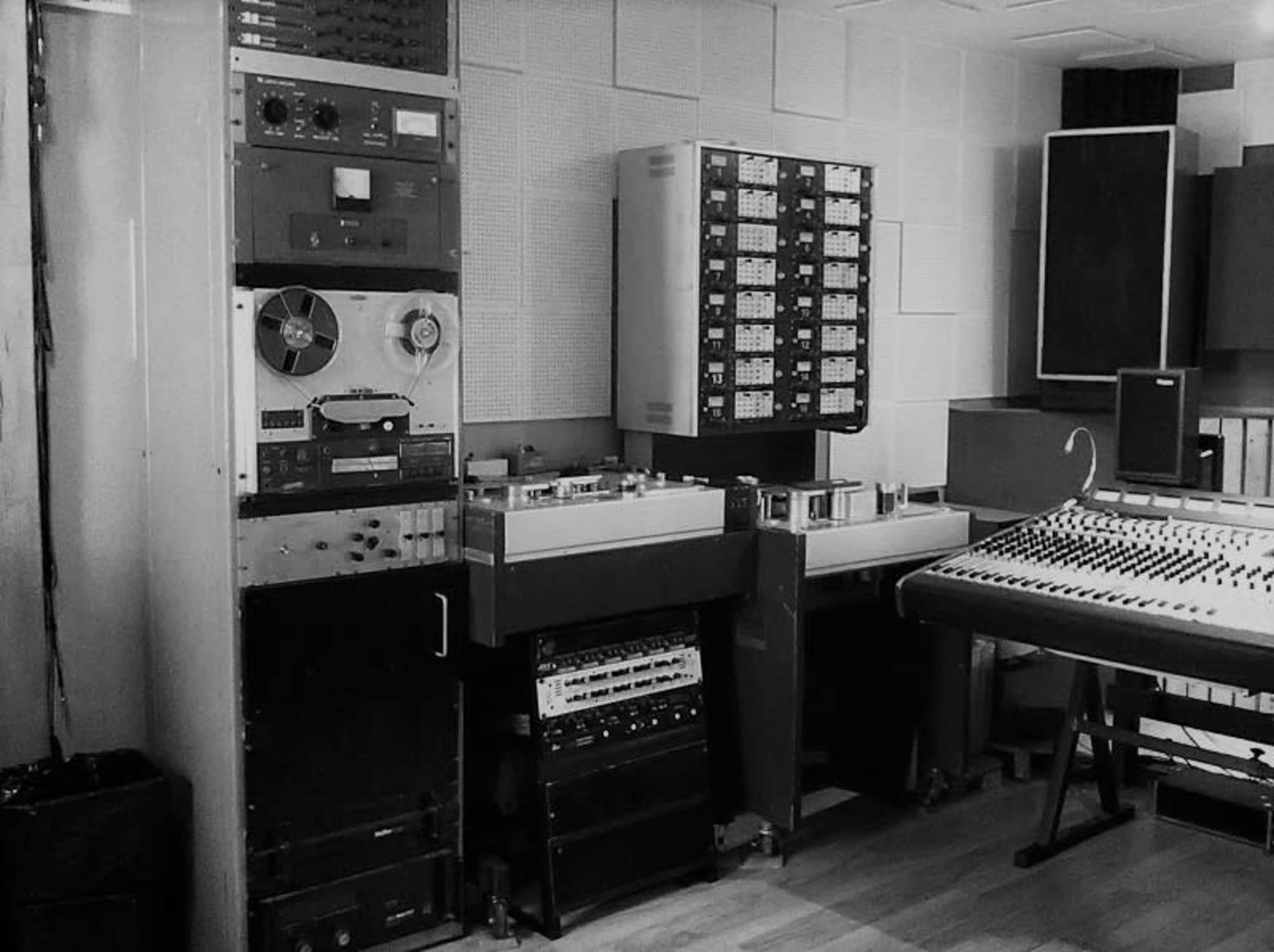 A recording studio with various audio equipment, including tape machines, a mixing console, and multiple racks of sound processors.