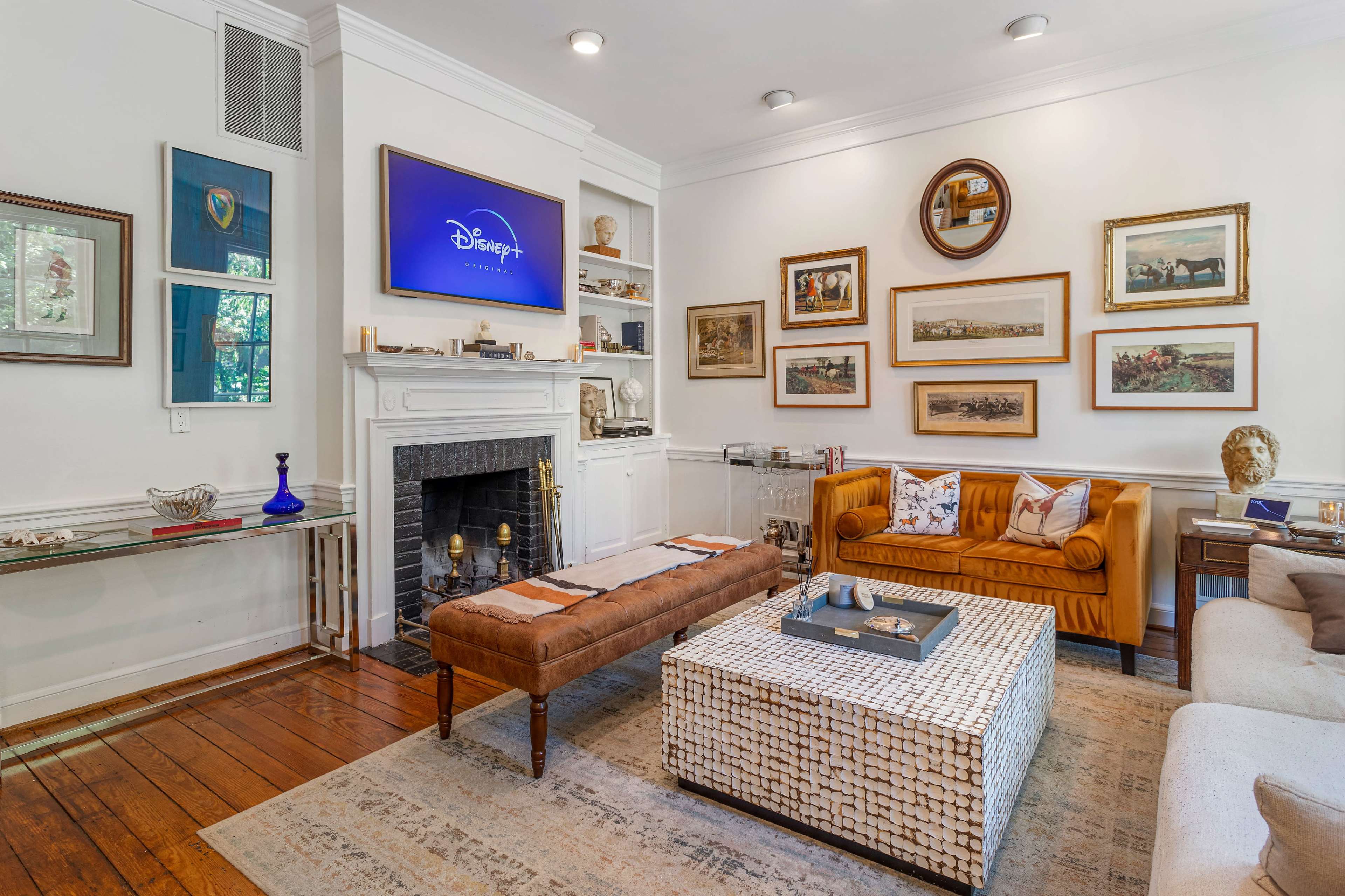 The living room features a mix of modern and classic decor, including a television above the fireplace, framed art on the walls, and a mix of seating arrangements with a coffee table in the center.