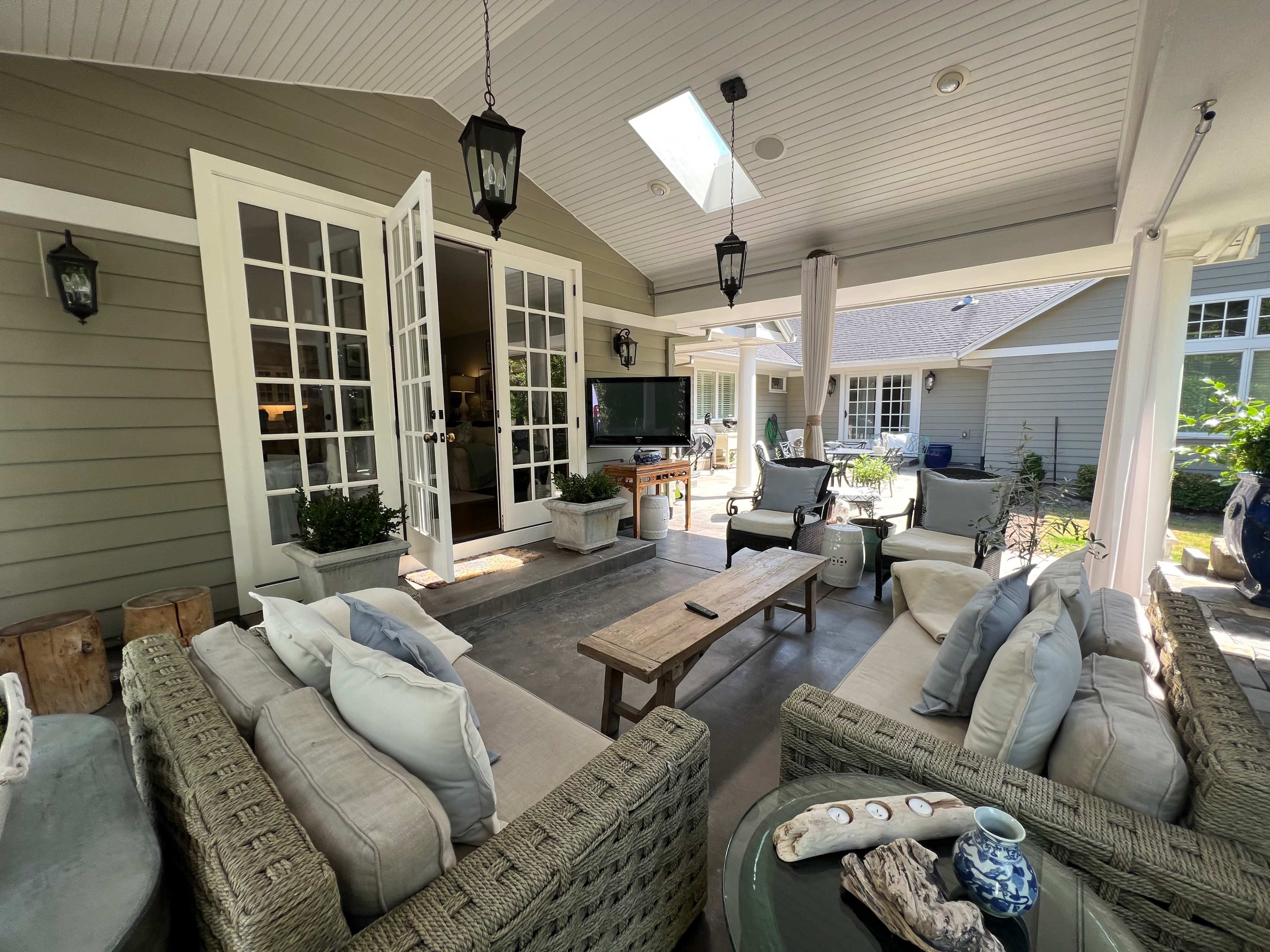 The image shows a covered outdoor living area with a sofa set, a coffee table, and a television mounted on the wall, surrounded by doors leading to other rooms.