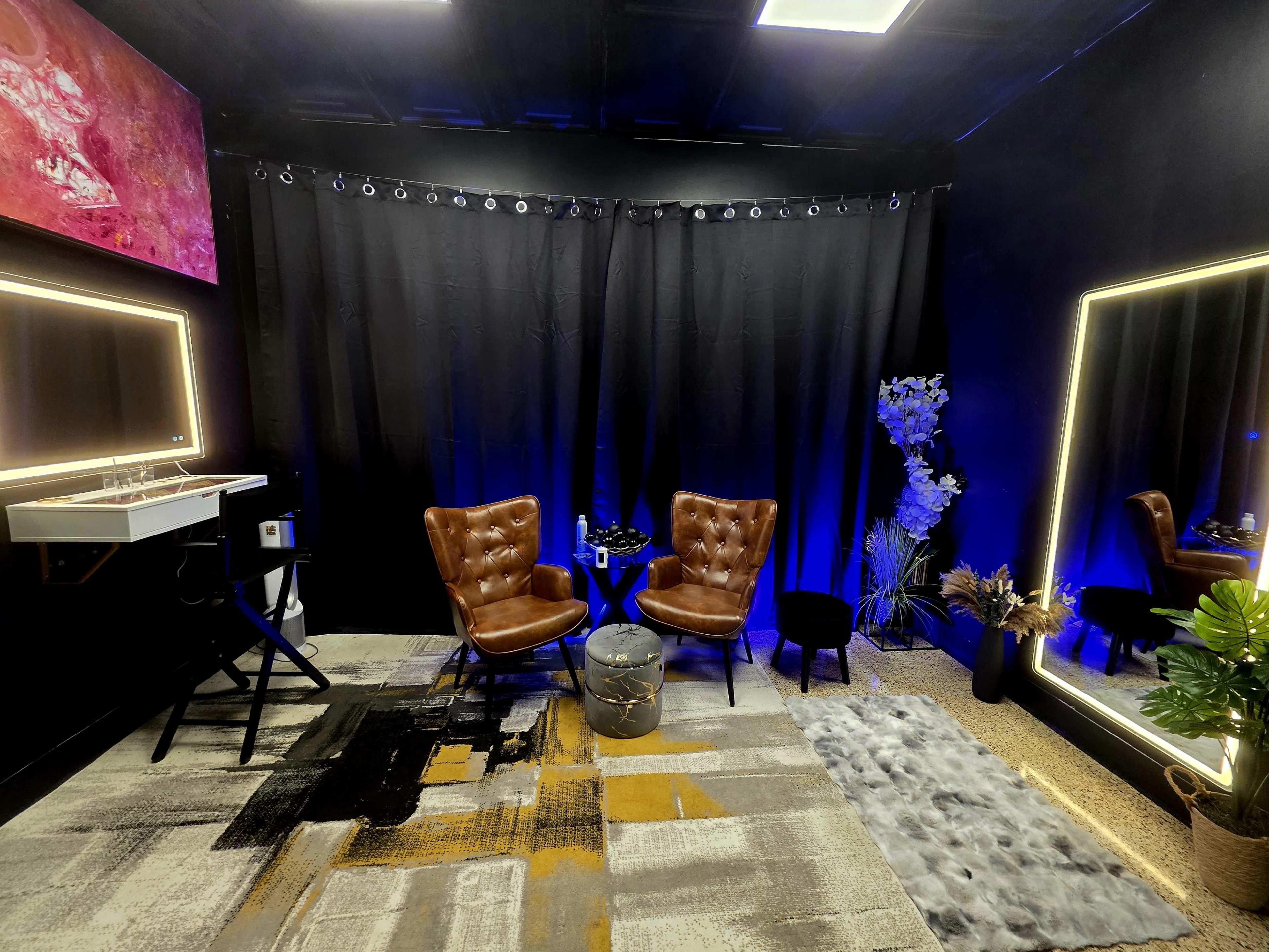 The image shows a modern room with two brown leather chairs, a small round table between them, and a large mirror, all set against a backdrop of black curtains and colorful lighting.