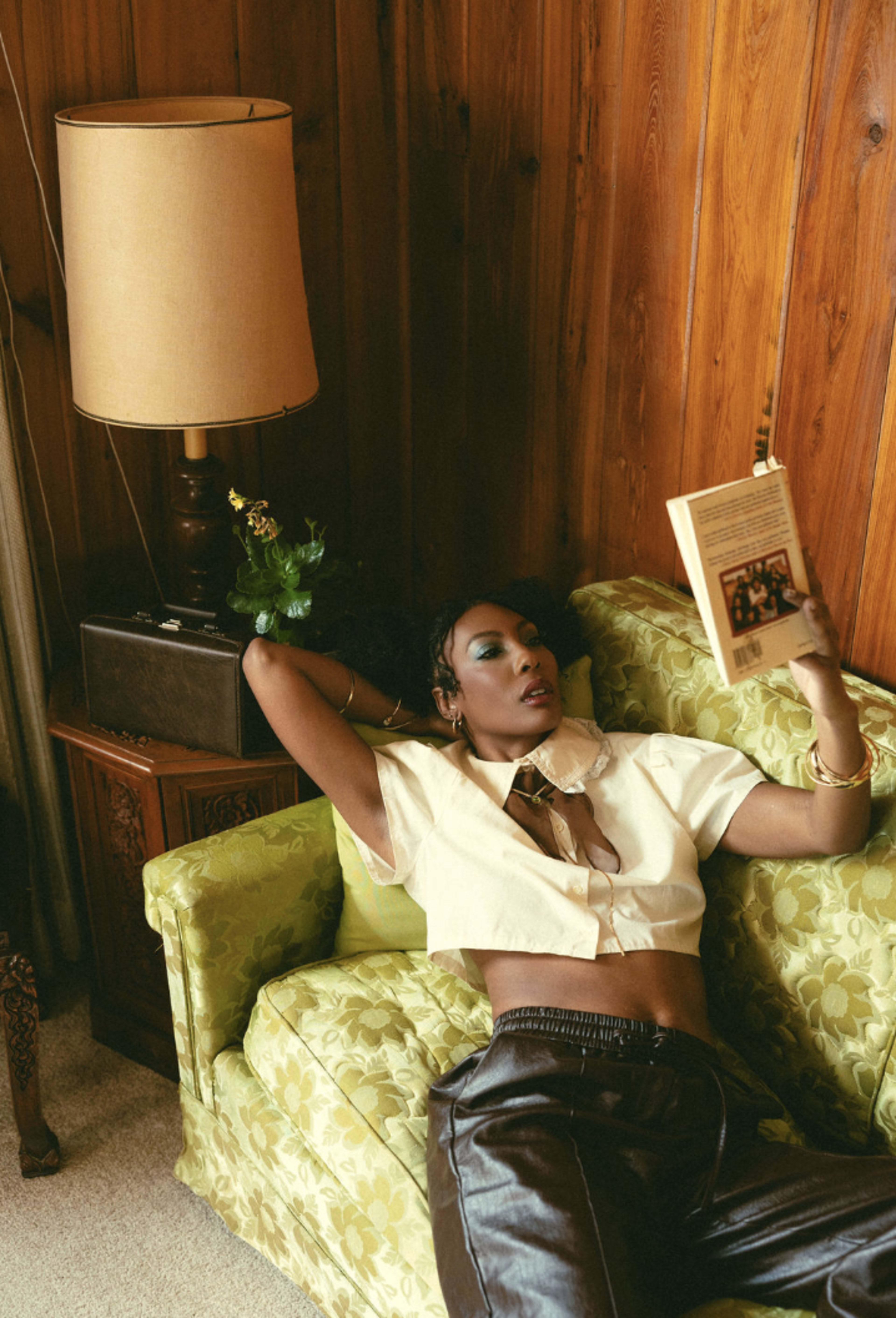 A person reclines on a green floral sofa, reading a book beside a lamp and a small decorative item.