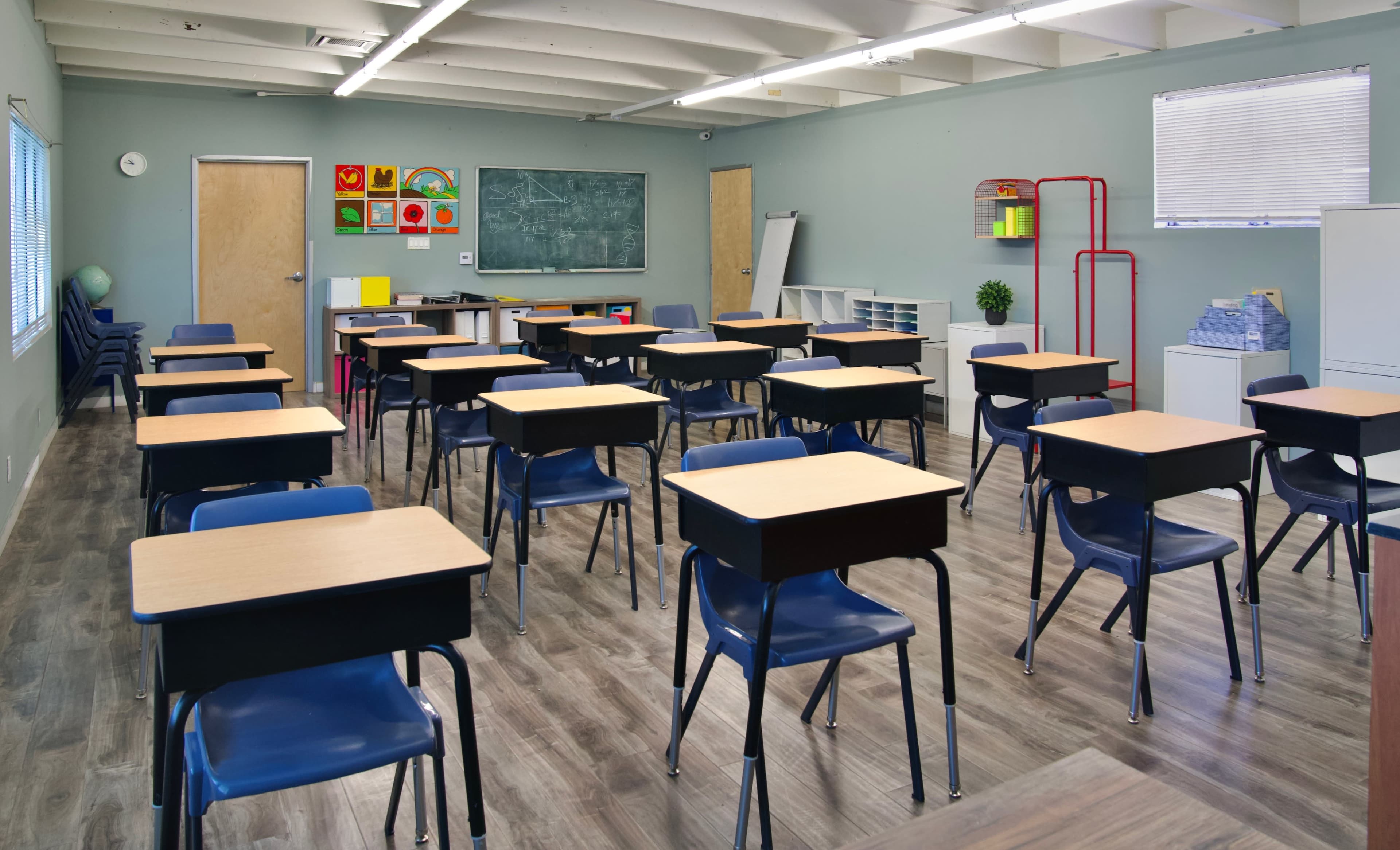 A classroom is set up with rows of desks and chairs, featuring a chalkboard and storage units along the walls.