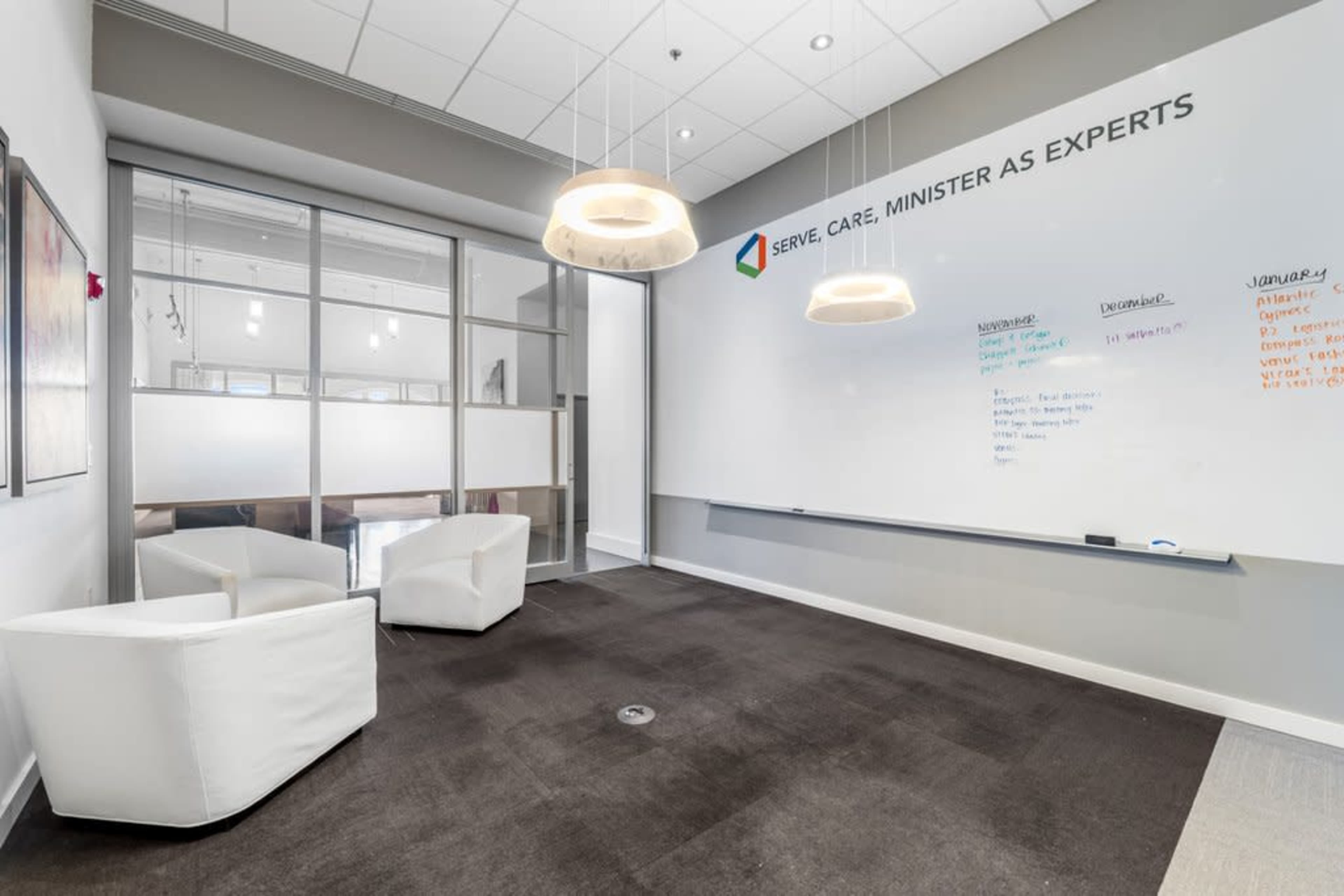 The image shows a modern office space featuring two white chairs, a large whiteboard with writing, and a glass partition in the background.