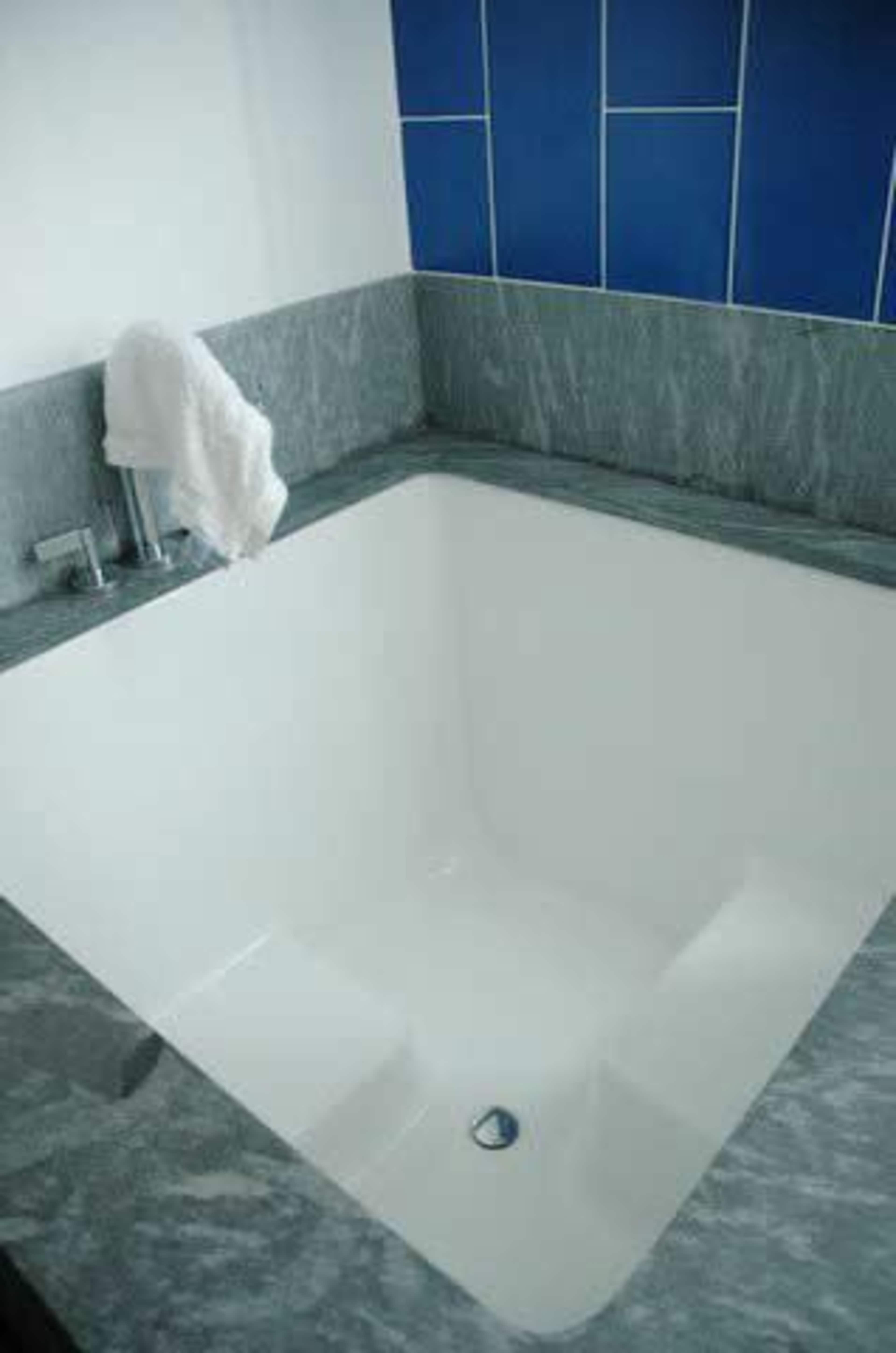 A square bathtub with a sleek white finish and marble surrounding, positioned against a wall with blue tiles.