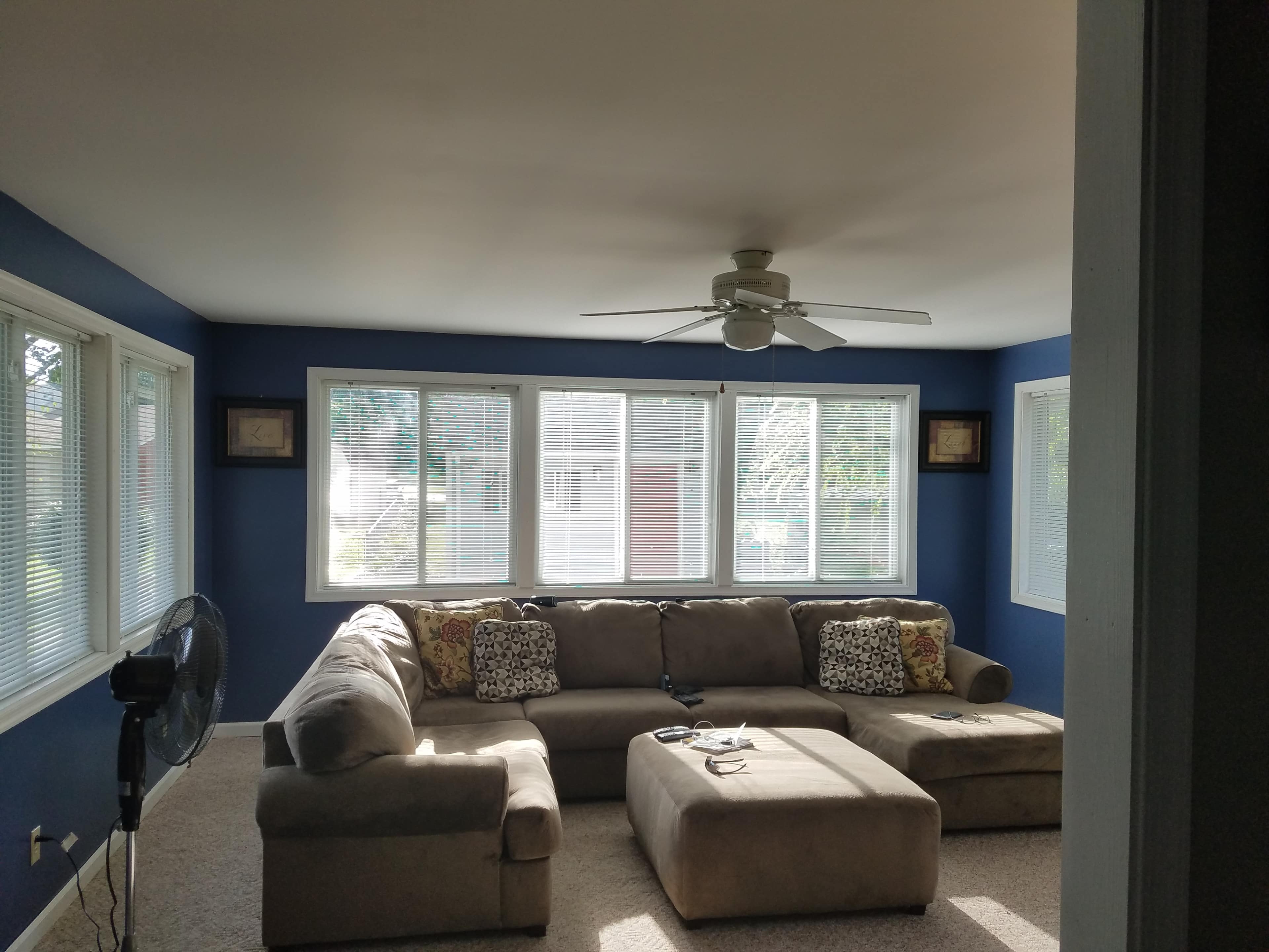 A living room features a large sectional sofa, an ottoman, and multiple windows allowing natural light to enter.