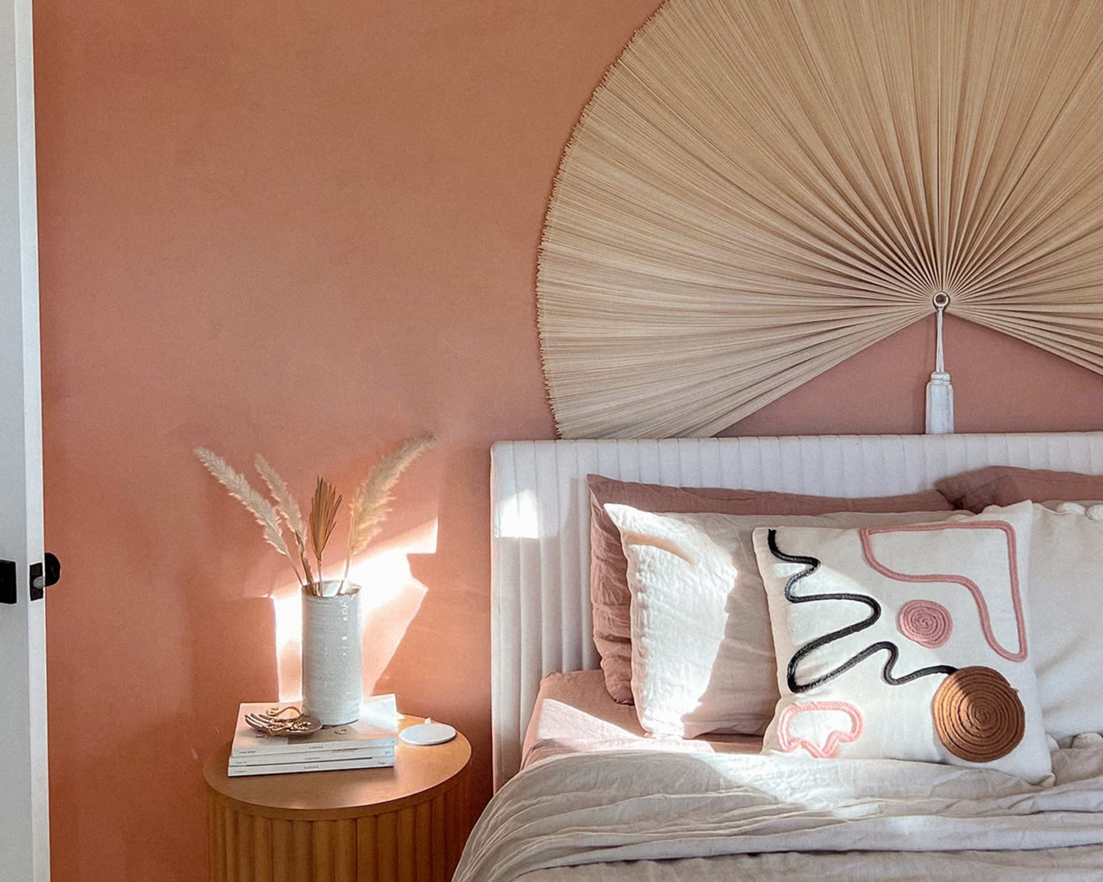 A neatly arranged bedroom features a pink wall, a large fan-shaped wall decoration above a bed with light-colored linens, and a circular nightstand holding decorative items and a vase with dried plants.