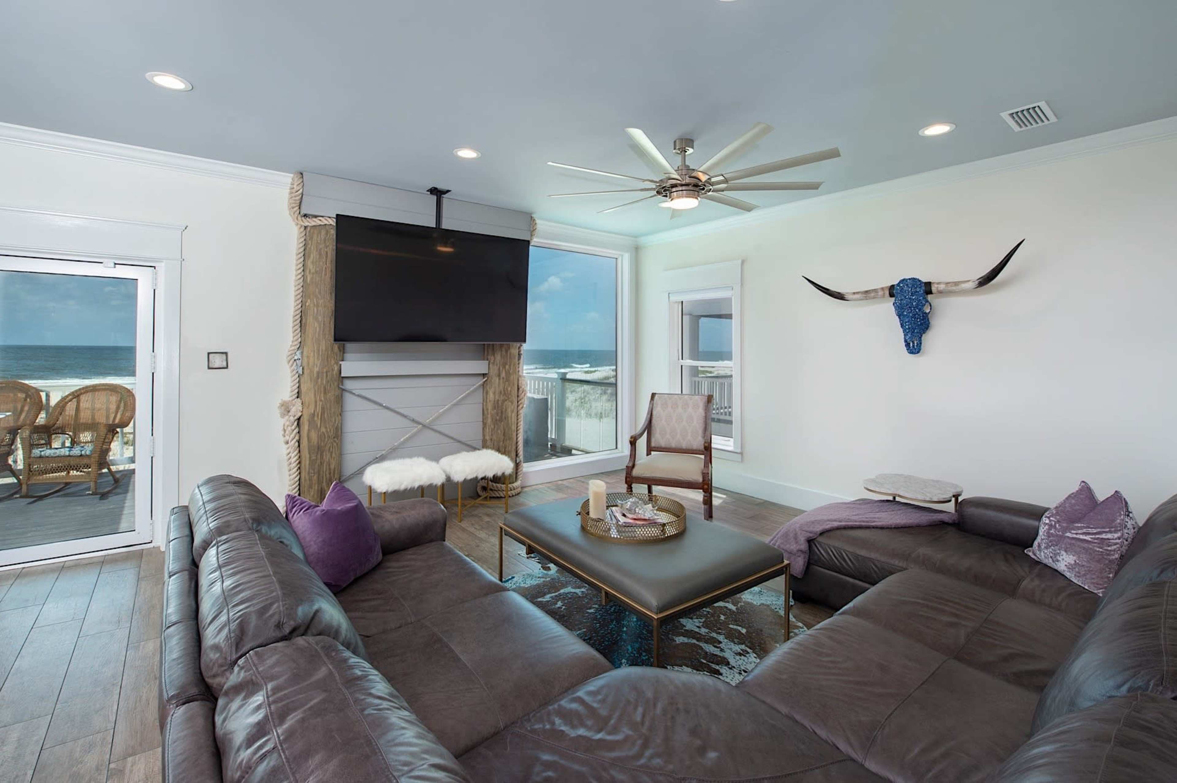 The living room features a large sectional sofa, a coffee table centered underneath a ceiling fan, and a television mounted above a decorative fireplace, with a view of the ocean through large windows.