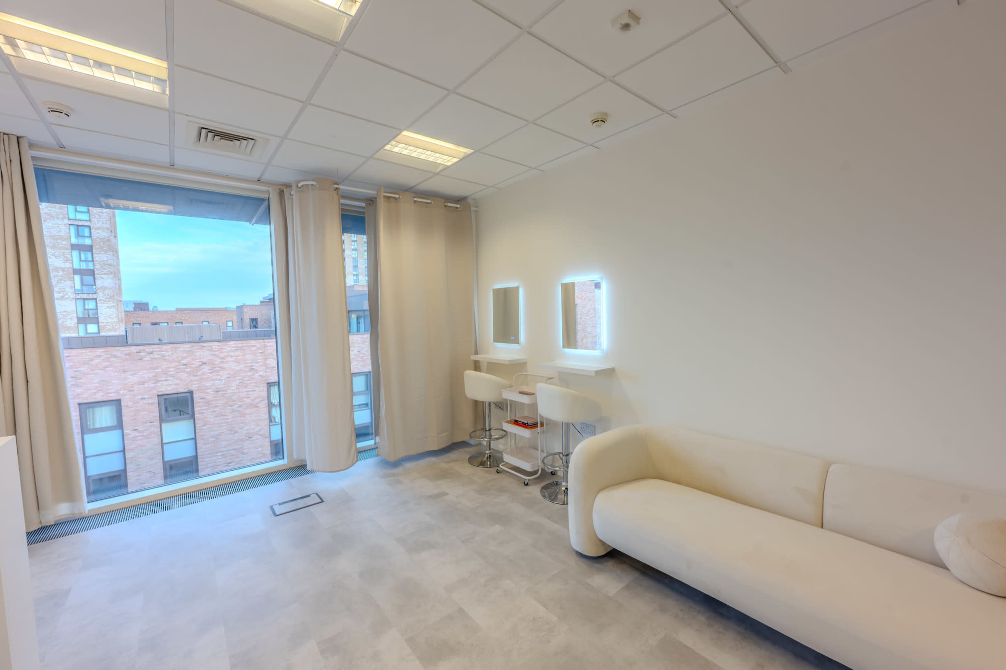 The image shows a minimalistic room with a white couch, two illuminated mirrors, and large windows revealing a brick building outside.