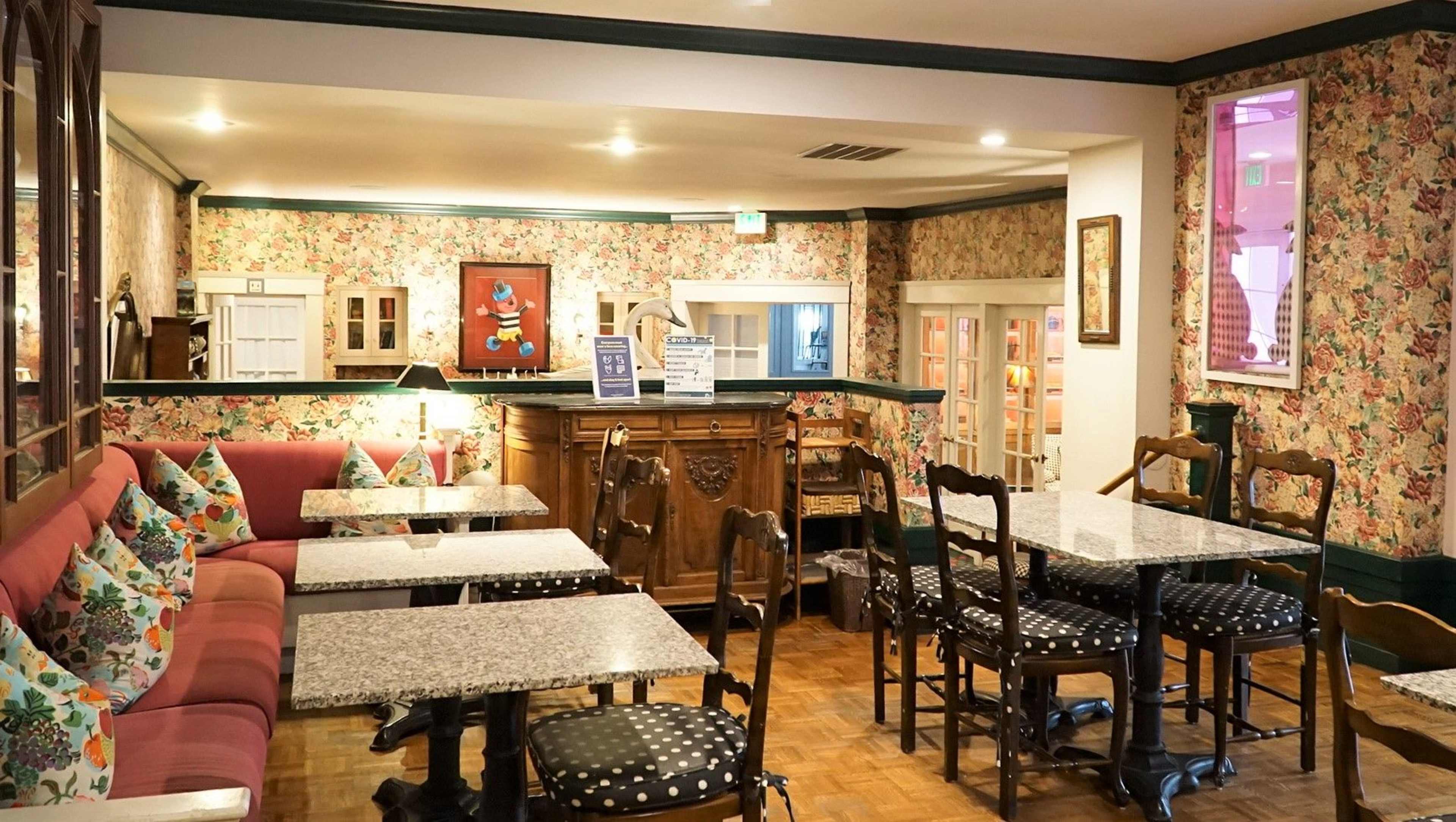 The image shows a quaint café interior featuring floral wallpaper, granite tabletops, wooden chairs, and a cozy seating area with cushions.