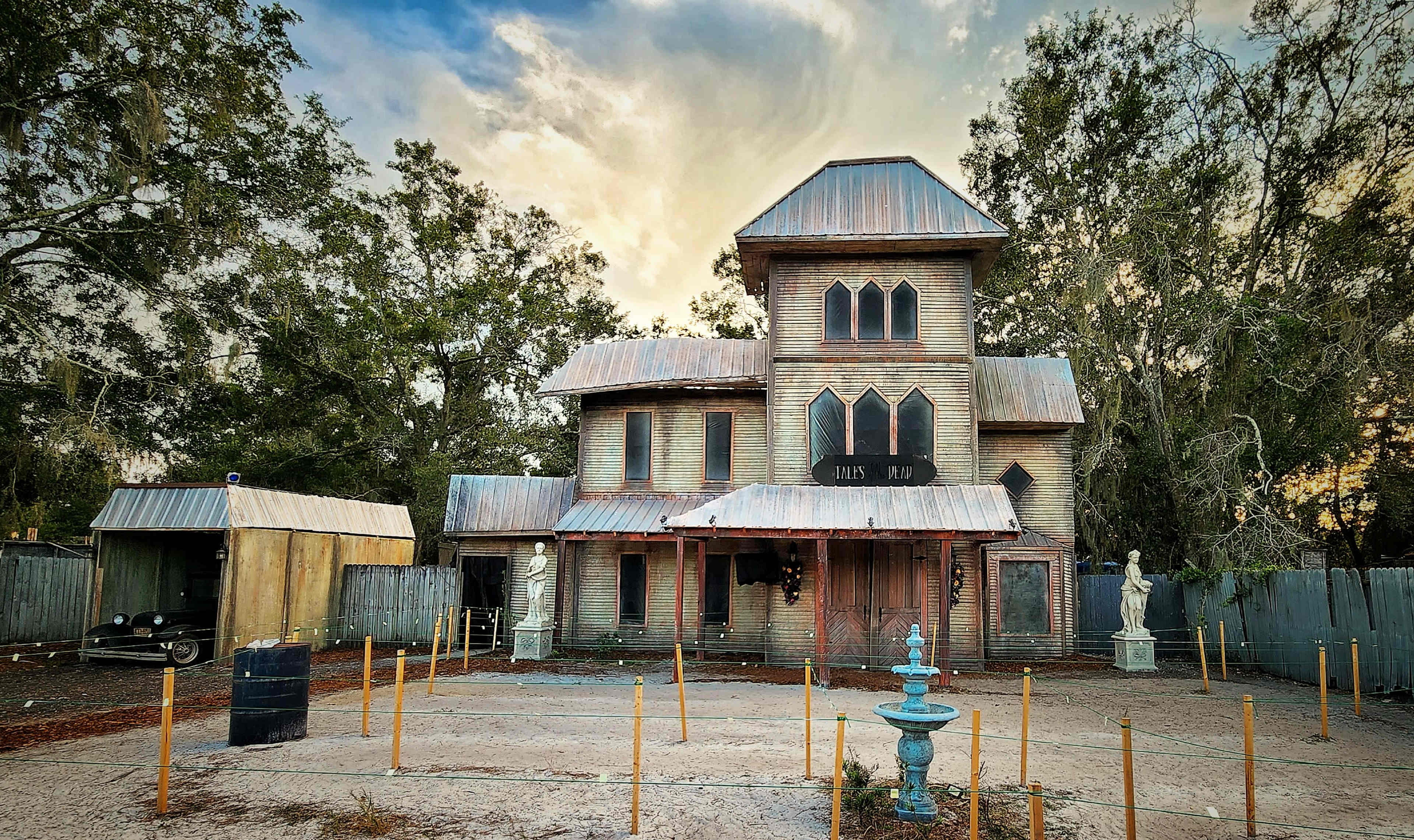 An old, dilapidated house with a metal roof stands among trees, featuring a front porch and an ornate fountain in the yard.