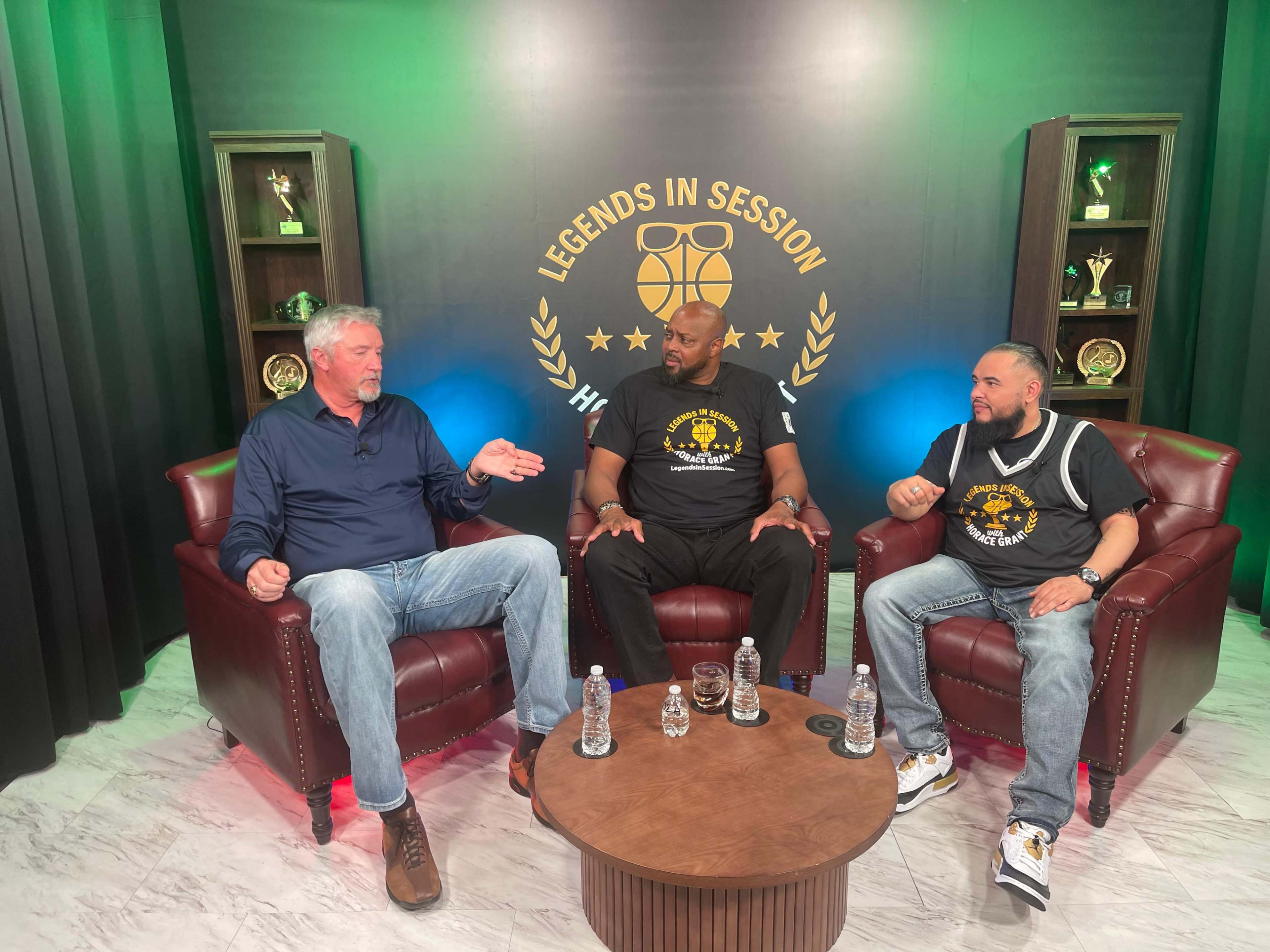 Three men are seated in armchairs in front of a backdrop featuring awards and the text "Legends in Session," engaged in a conversation.