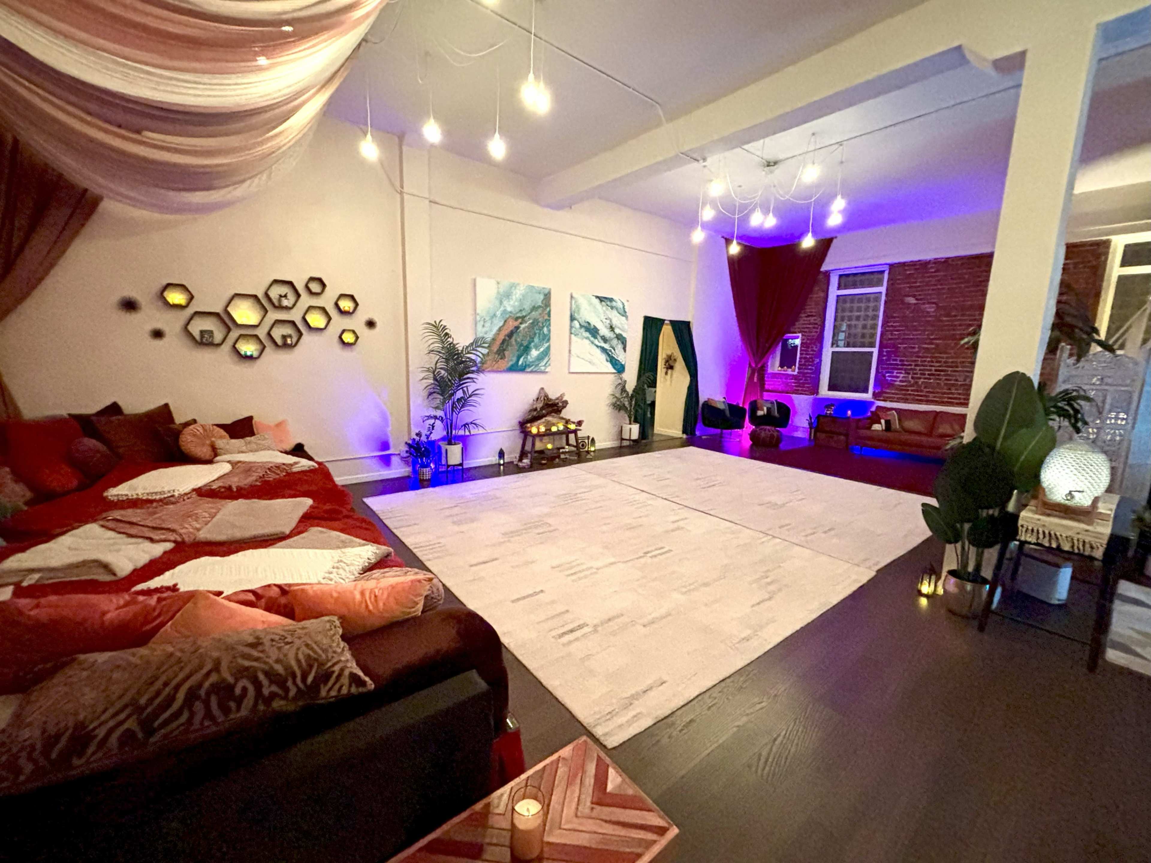 A spacious, well-lit room features a mix of decorative elements, including a large area rug, comfortable seating, and ambient lighting with draped fabrics overhead.
