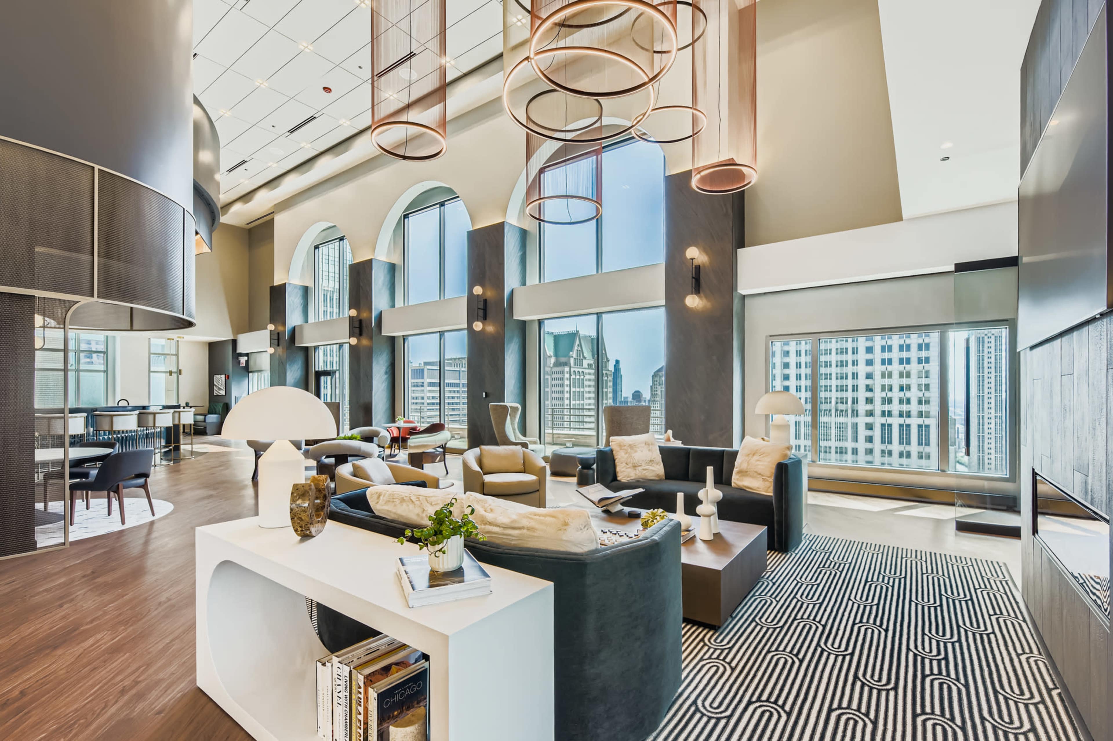 The image shows a modern lounge area with large windows, stylish furniture, and circular light fixtures in an upscale setting.