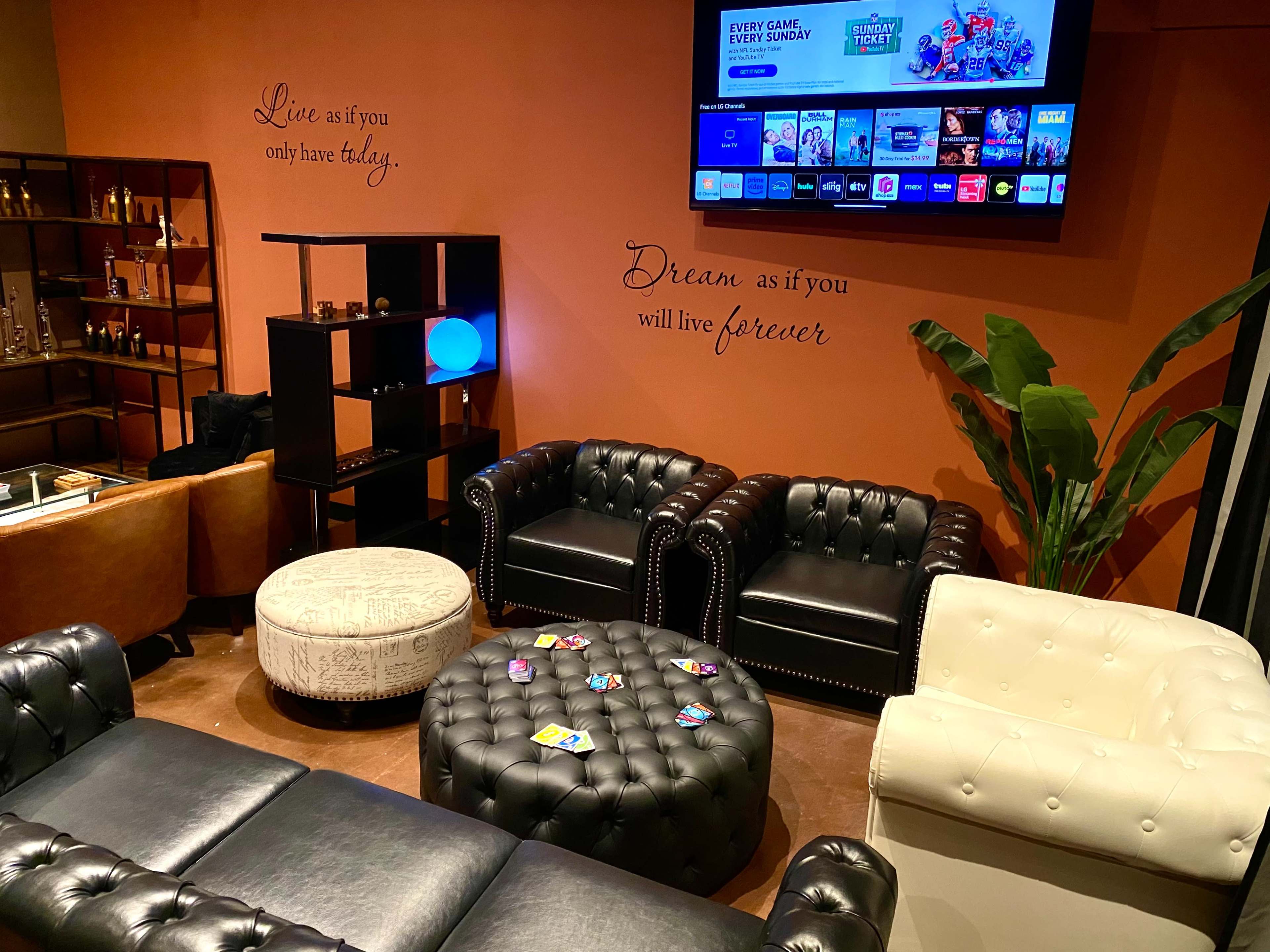 The image shows a cozy lounge area with black leather chairs, a white sofa, an ottoman with colorful items on it, and a wall featuring motivational phrases.